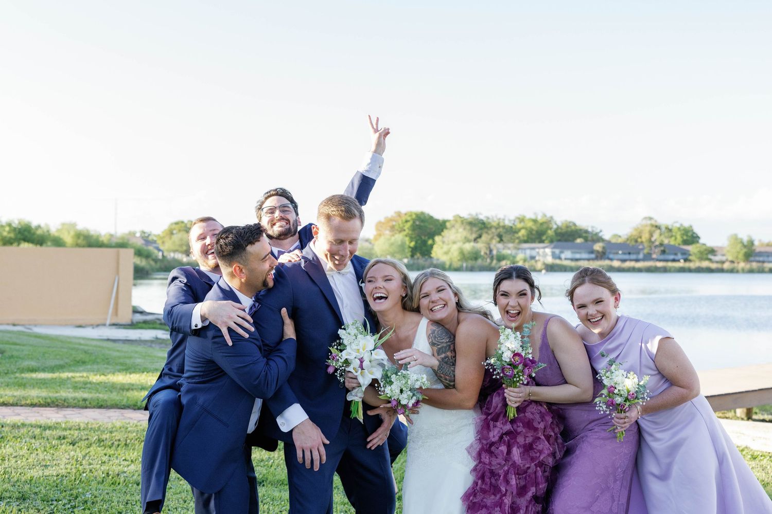 Bride and groom celebrate with wedding party on lawn near waterfront, bridesmaids in purple dresses.