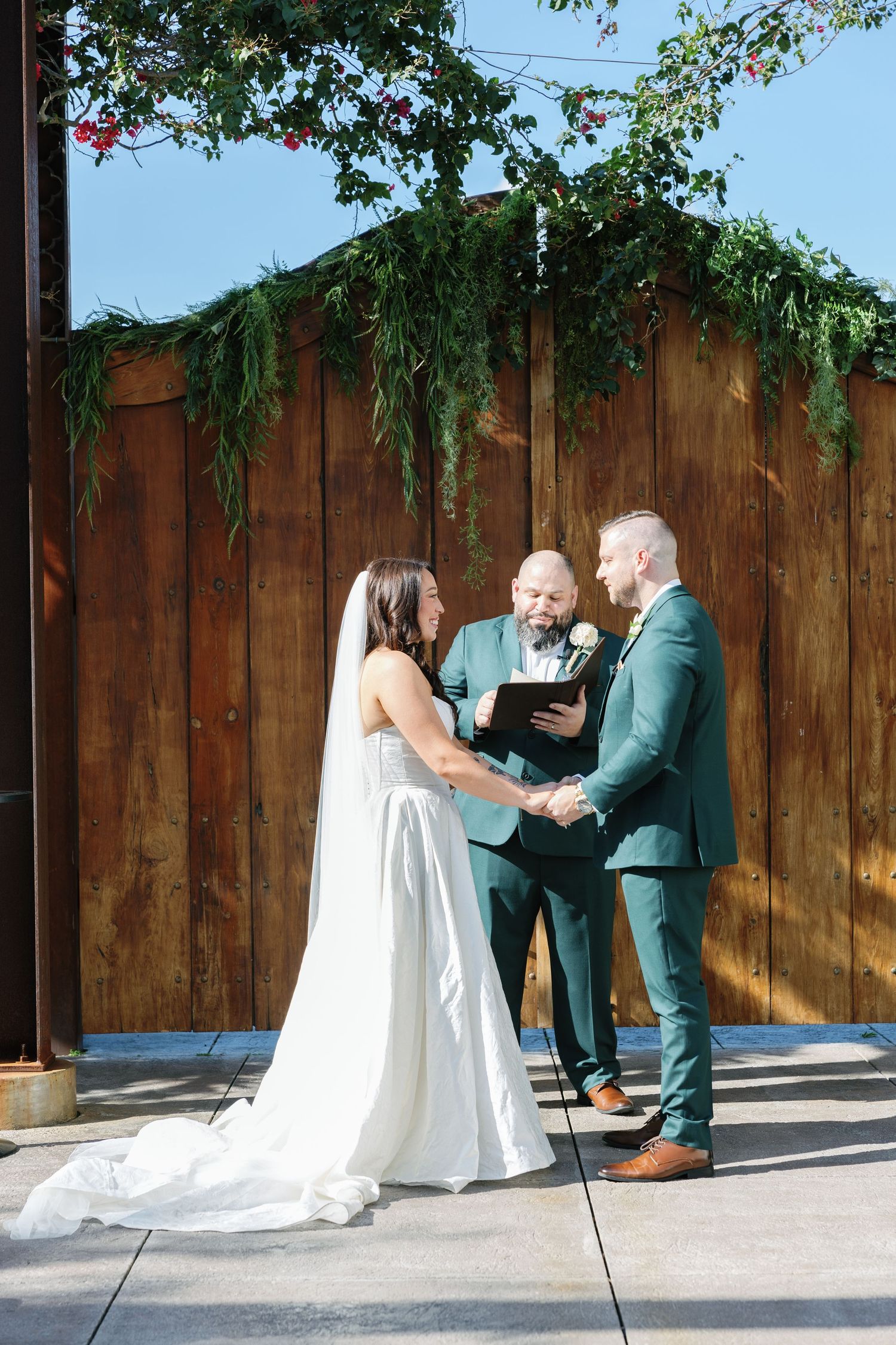 Bride and groom exchange vows outdoors before an officiant beneath a lush greenery arch on a sunny day.