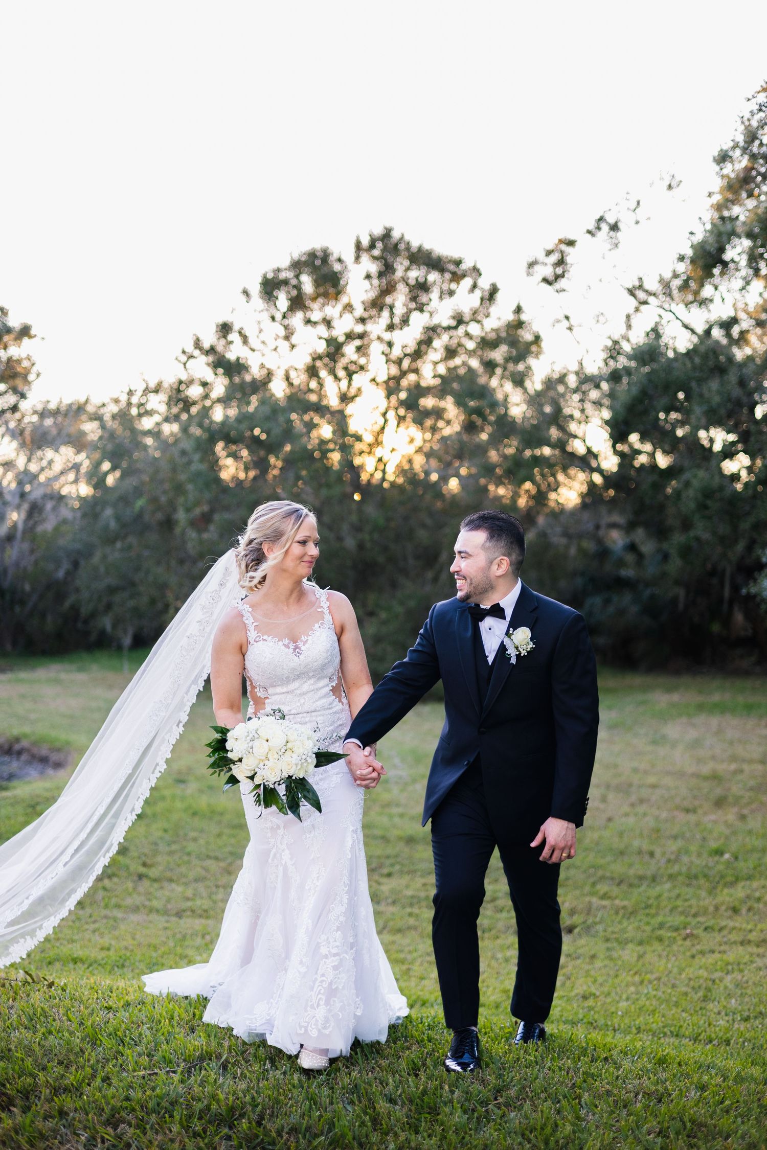 Bride and groom walk hand in hand through green lawn at golden hour sunset wedding.