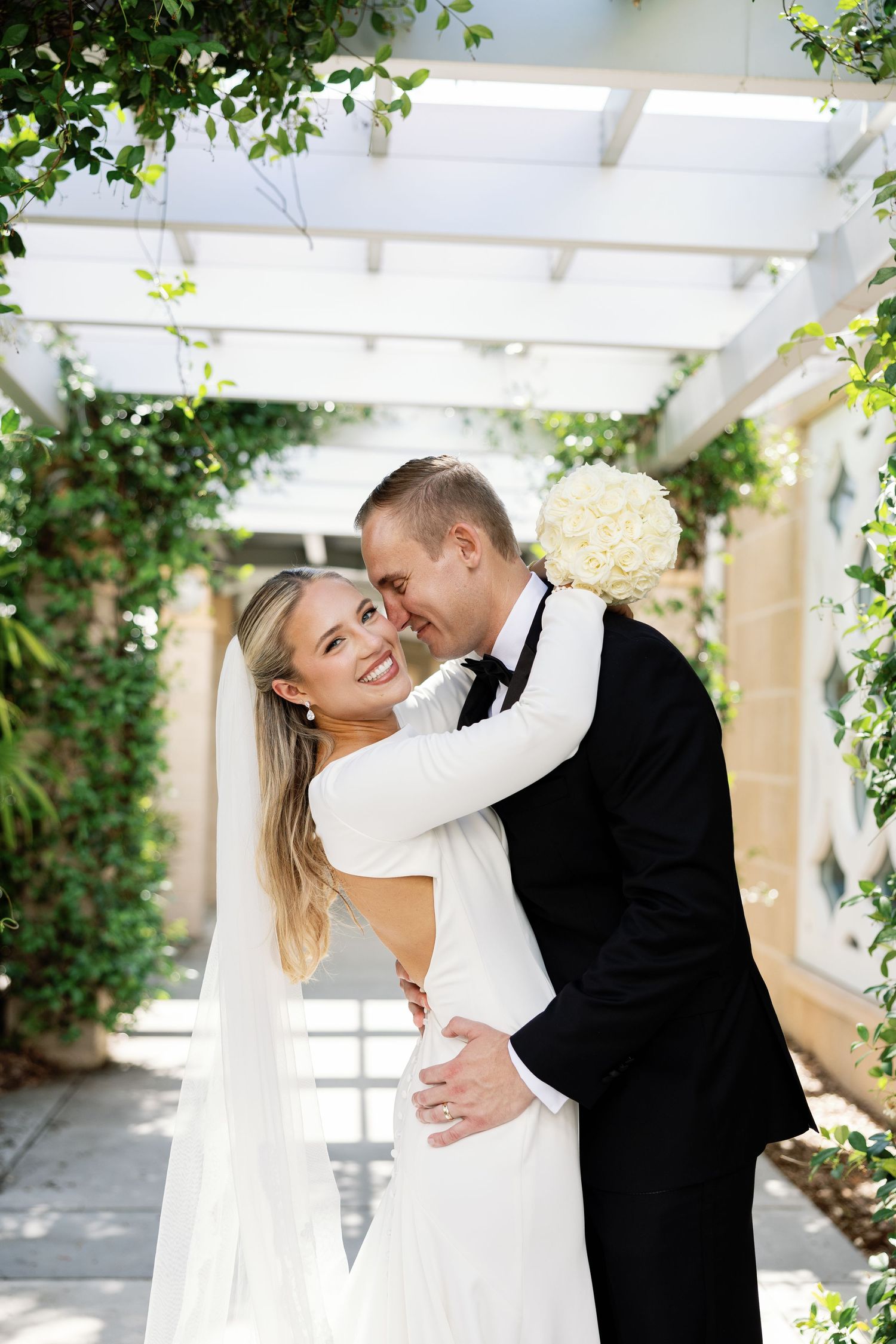 Smiling bride embraced by groom under vine-covered pergola in sunny outdoor wedding photo.