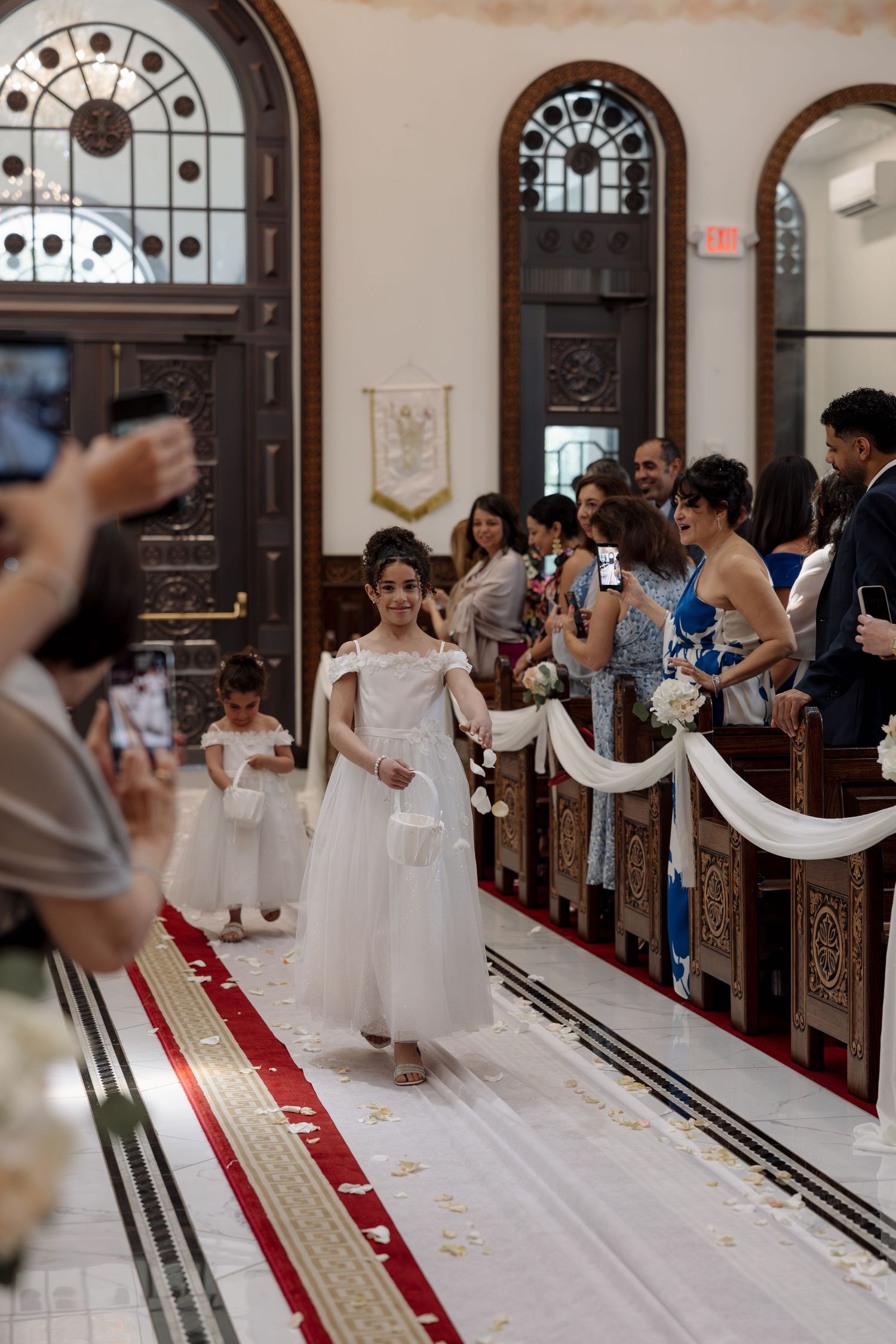 Young flower girl in white dress walks down church aisle during wedding ceremony while guests capture photos.