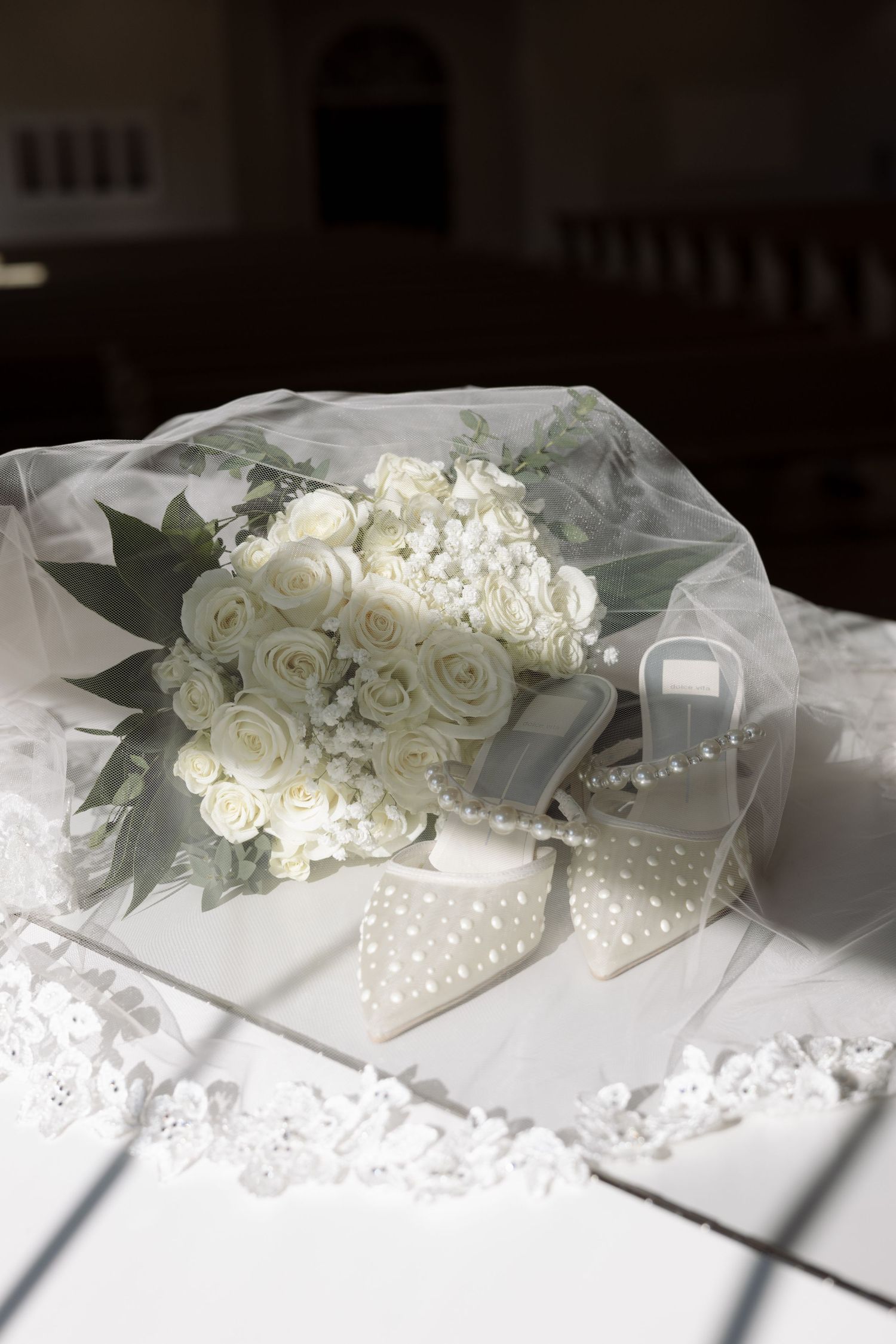 Elegant white rose bridal bouquet resting on lace veil beside small gift box in church setting.