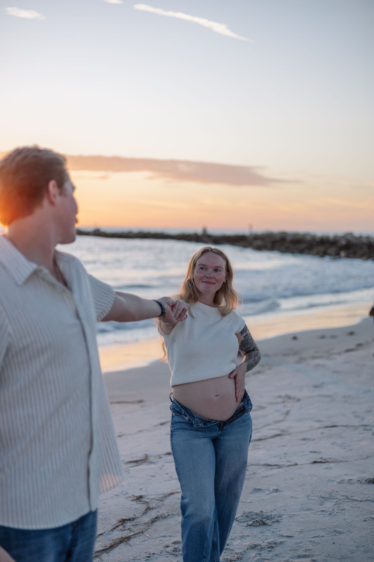 Pregnant woman with tattoos holding partner's hand on beach at sunset, smiling toward camera.