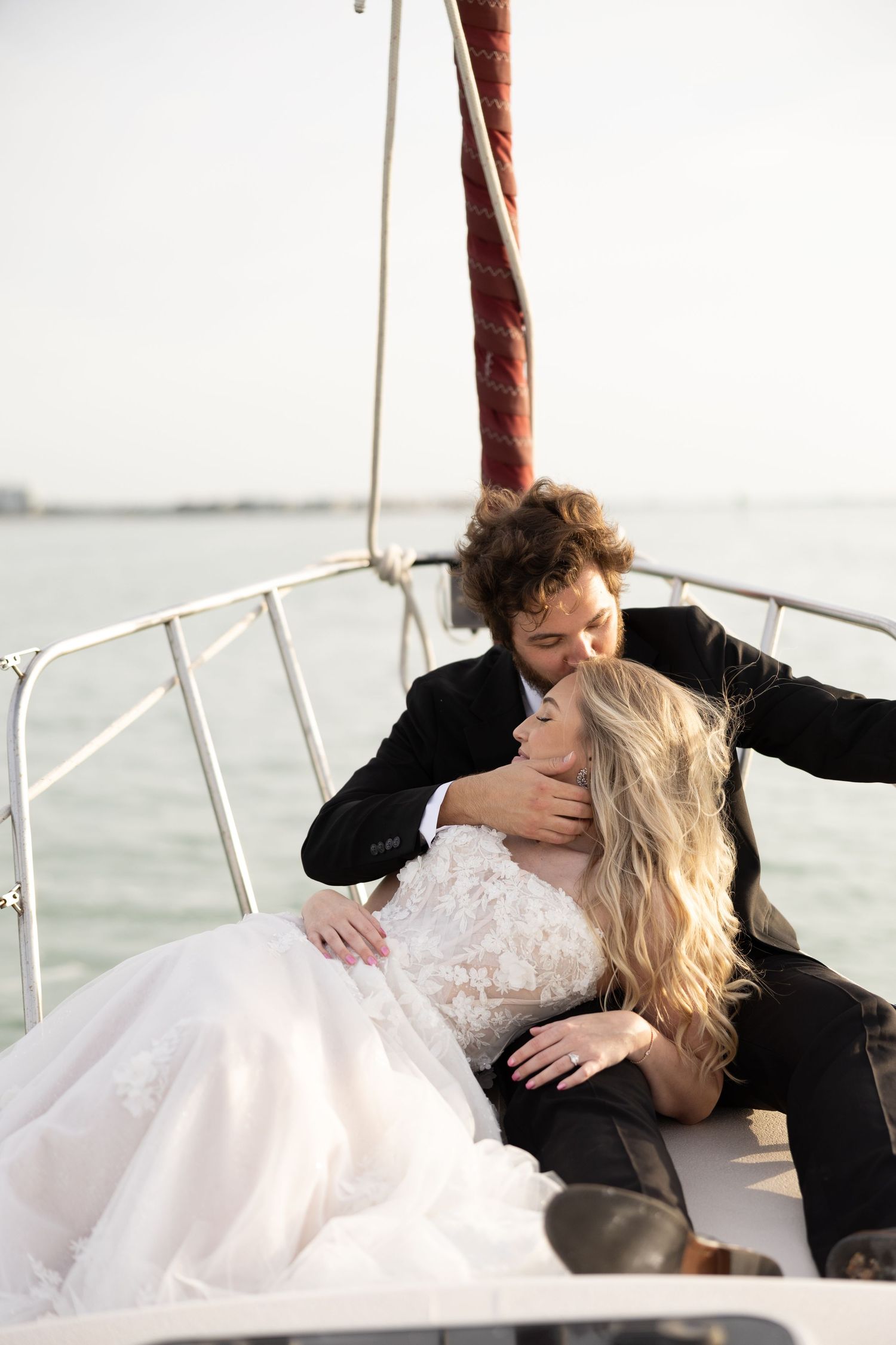 Bride and groom sharing romantic moment on sailboat, bride in lace gown, ocean backdrop.