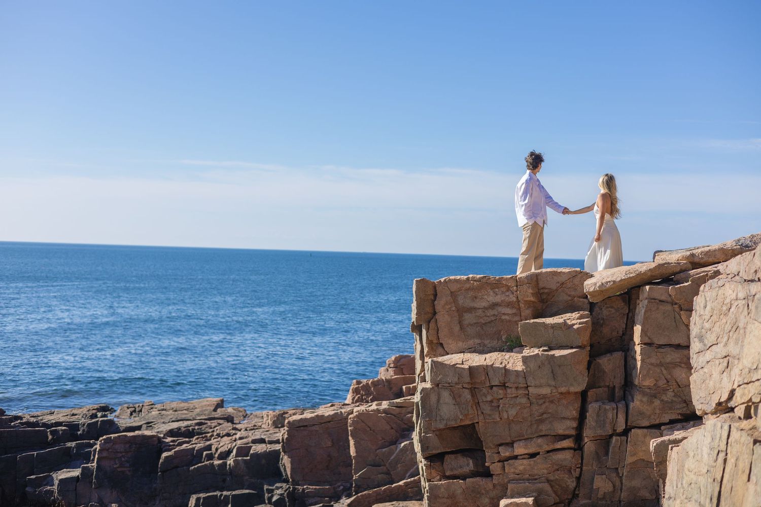 Romantic couple holding hands on dramatic rocky ocean cliffs under bright blue sky at scenic coastal location.