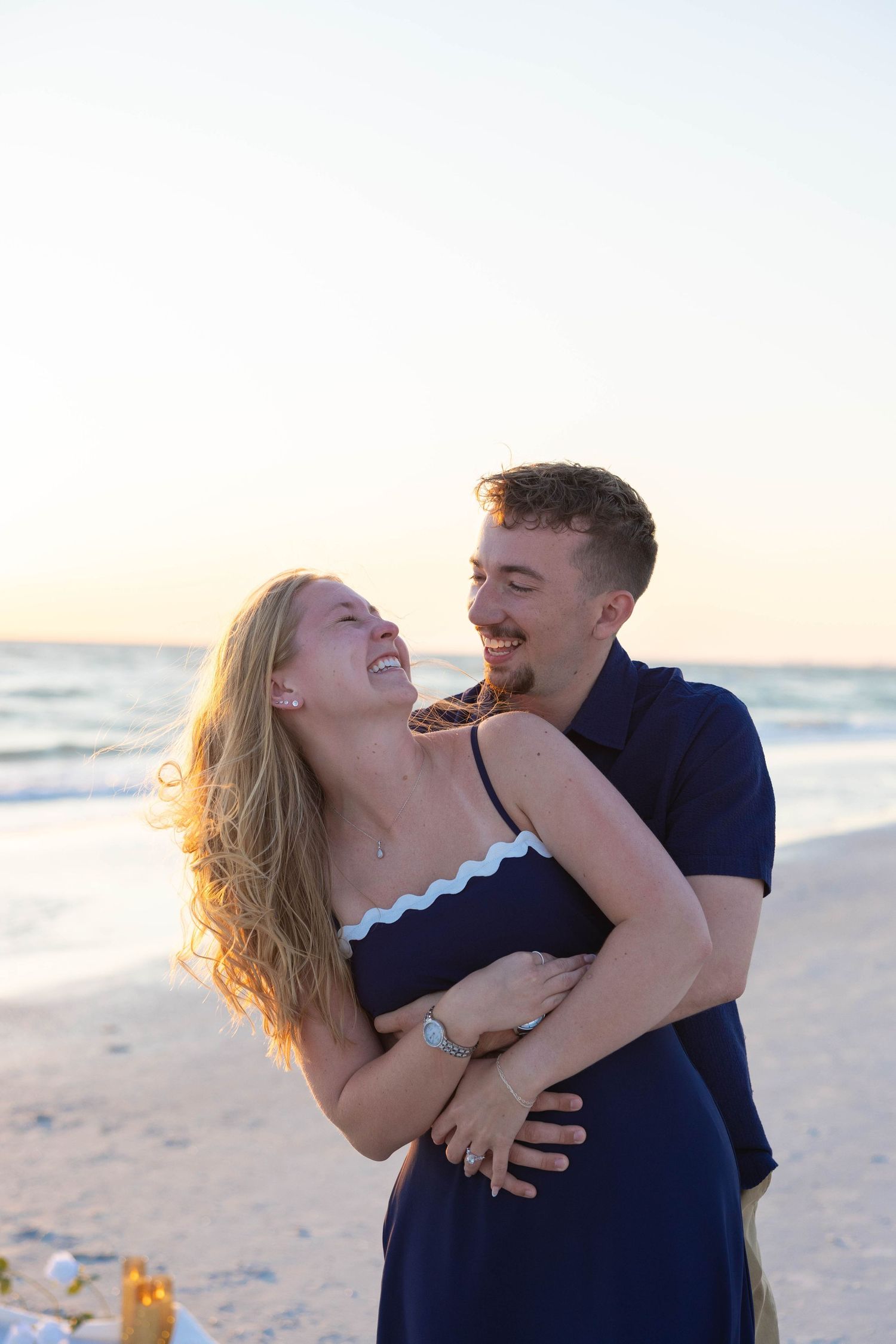 Newly engaged couple laughs and embraces joyfully on a beach during a golden sunset.