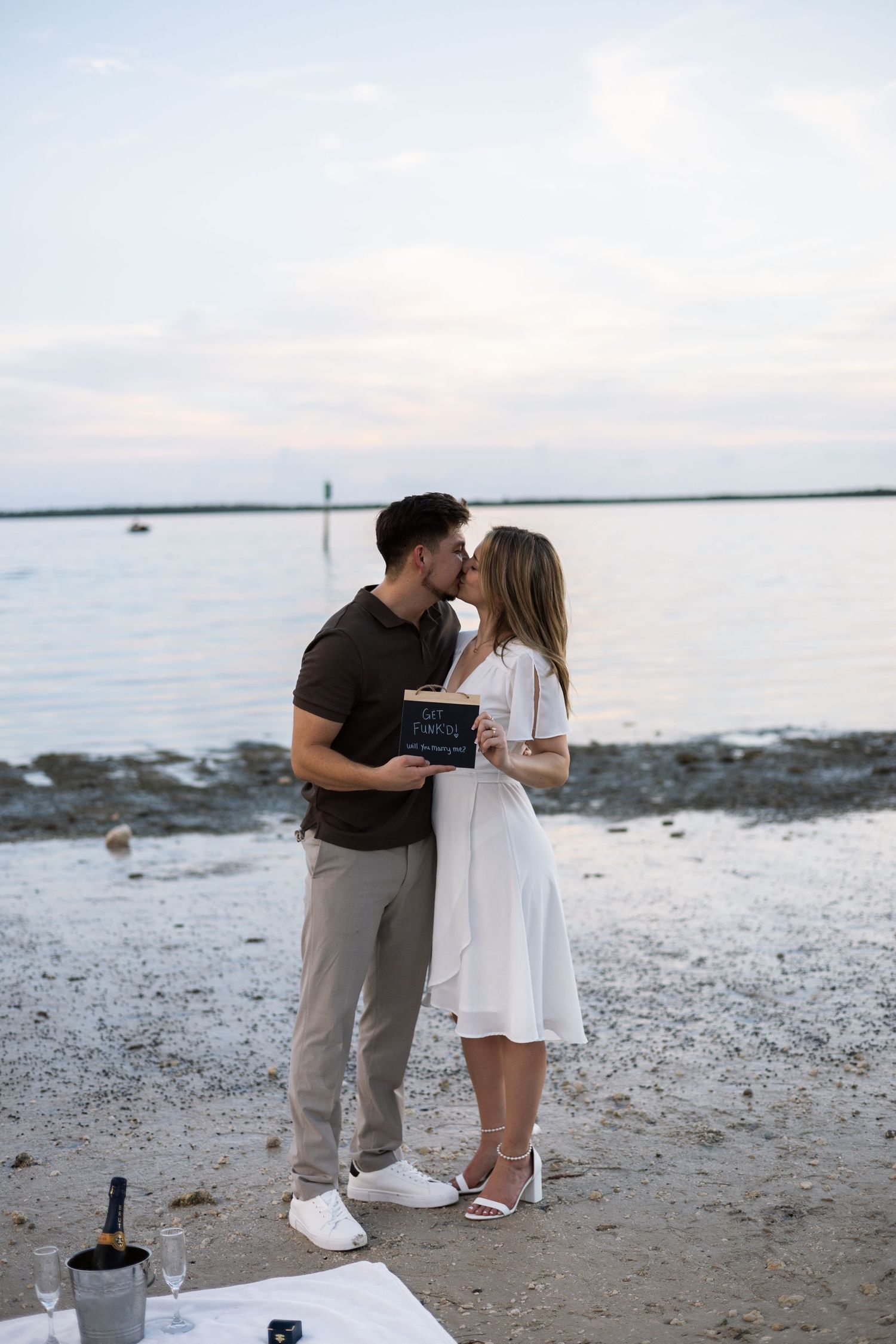 Newly engaged couple kissing on the beach holding a sign, with champagne bottle nearby at sunset.