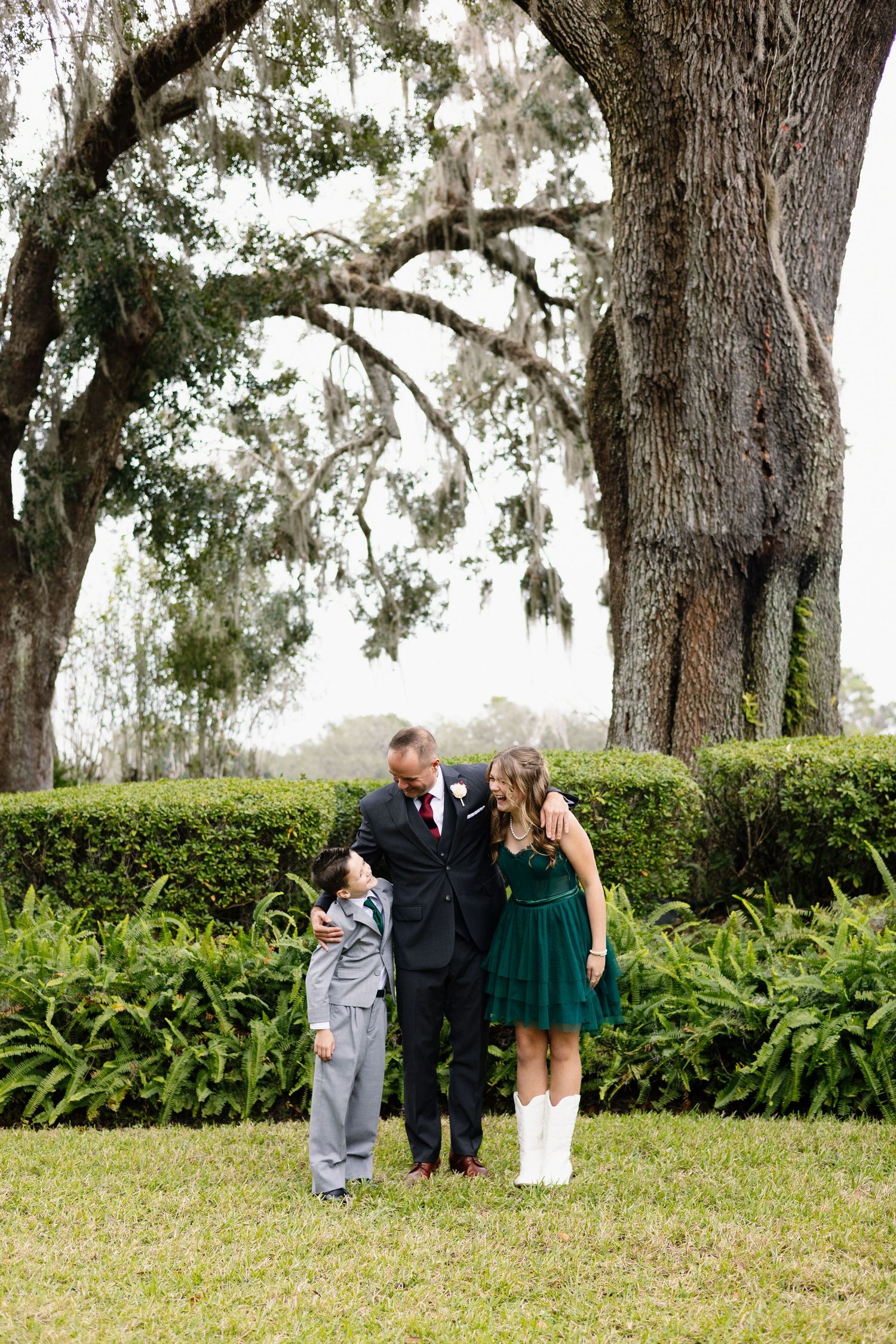 Groom in black suit with two children in formal attire standing under a large oak tree outdoors.