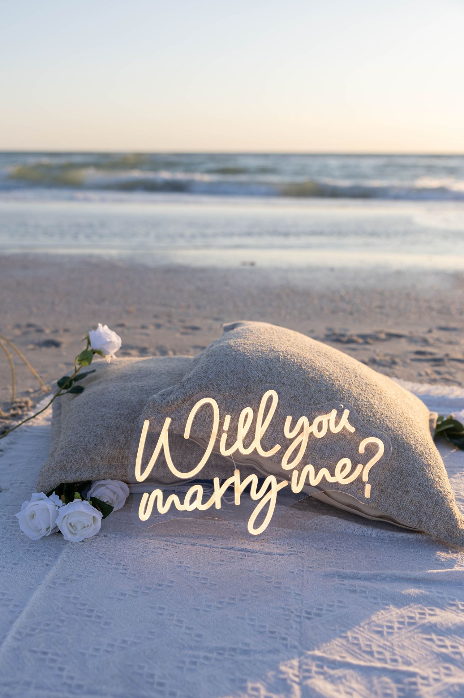 A glowing neon Will You Marry Me sign rests on a pillow surrounded by white roses on a serene beach at sunset.