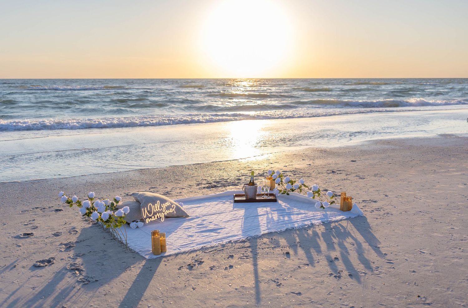An elegant beach picnic setup with white blanket, flowers, candles and lantern glowing at golden sunset.