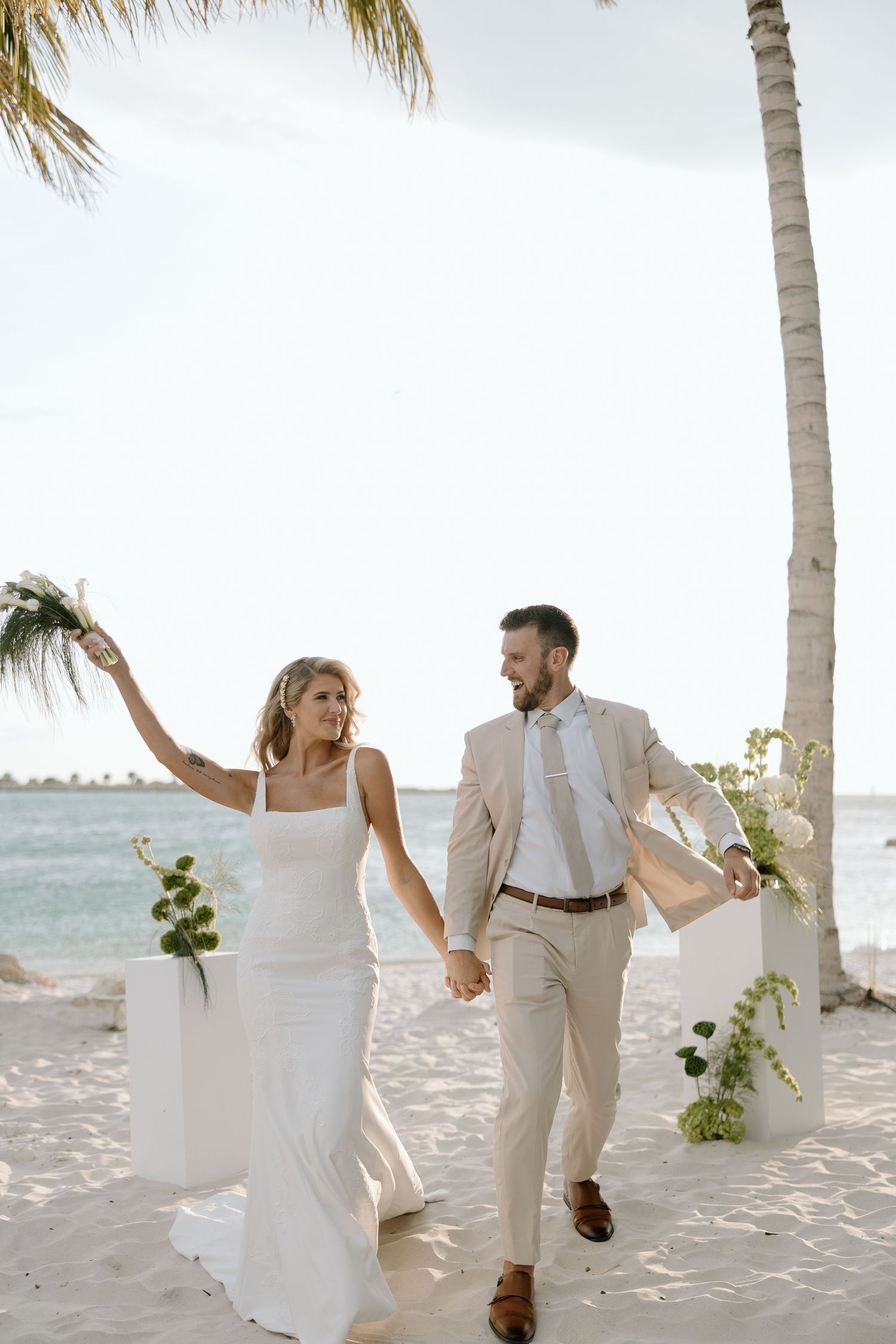 Smiling bride and groom walking hand in hand on tropical beach with white floral ceremony arch and palm trees.