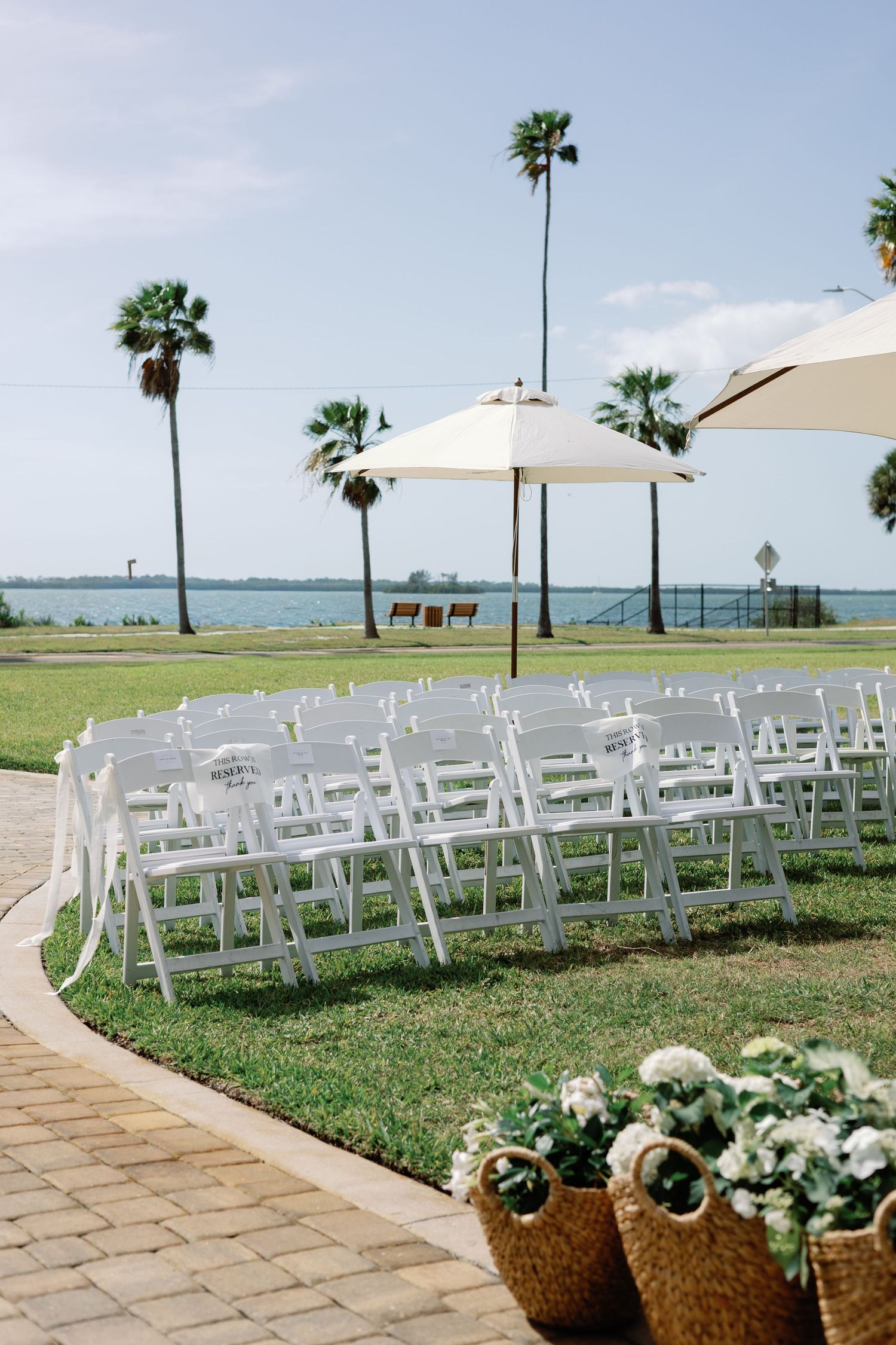 Outdoor waterfront wedding ceremony setup with white folding chairs, umbrellas, and palm trees by the bay.