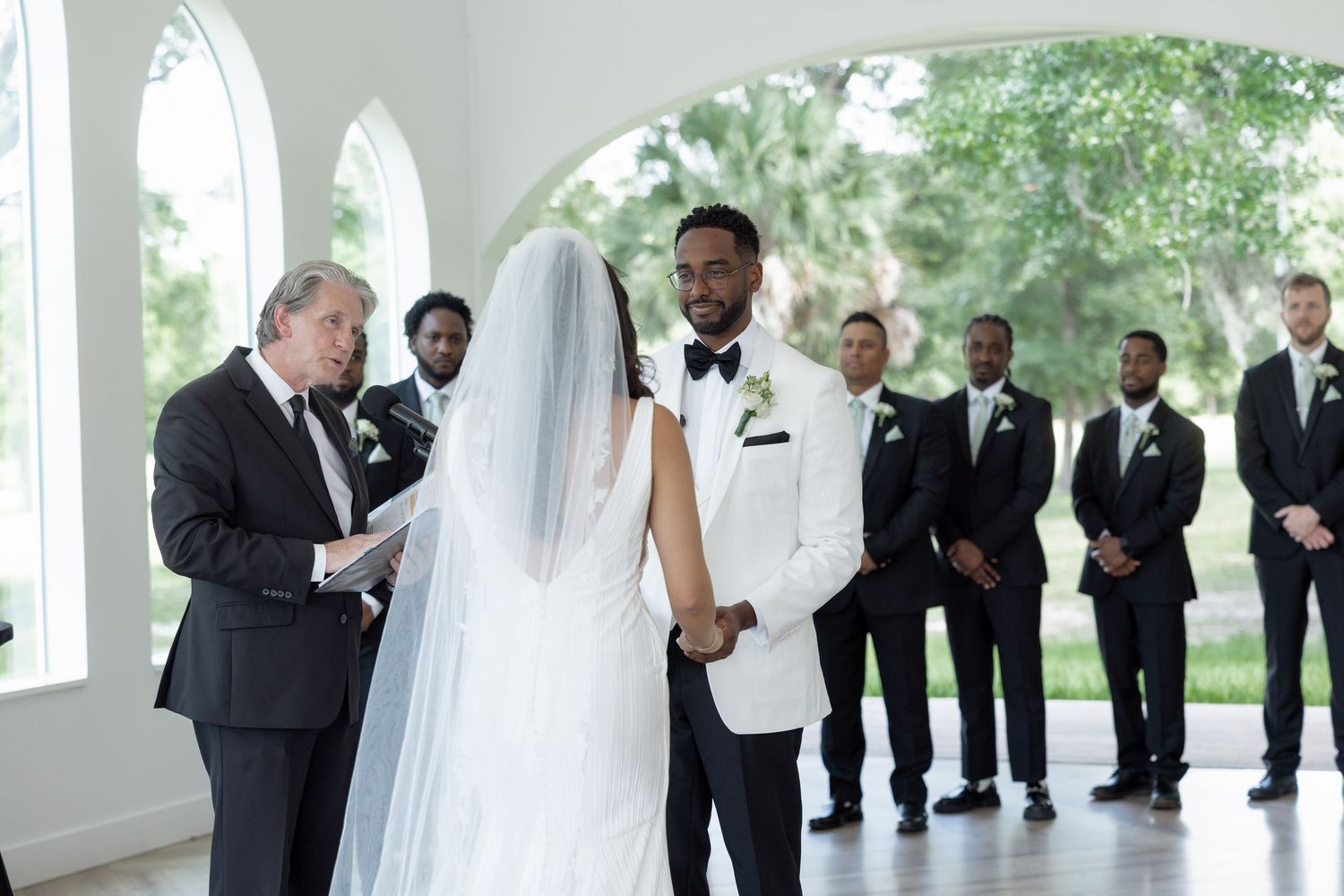 Groom in white tuxedo holds brides hands as officiant reads vows during wedding ceremony.