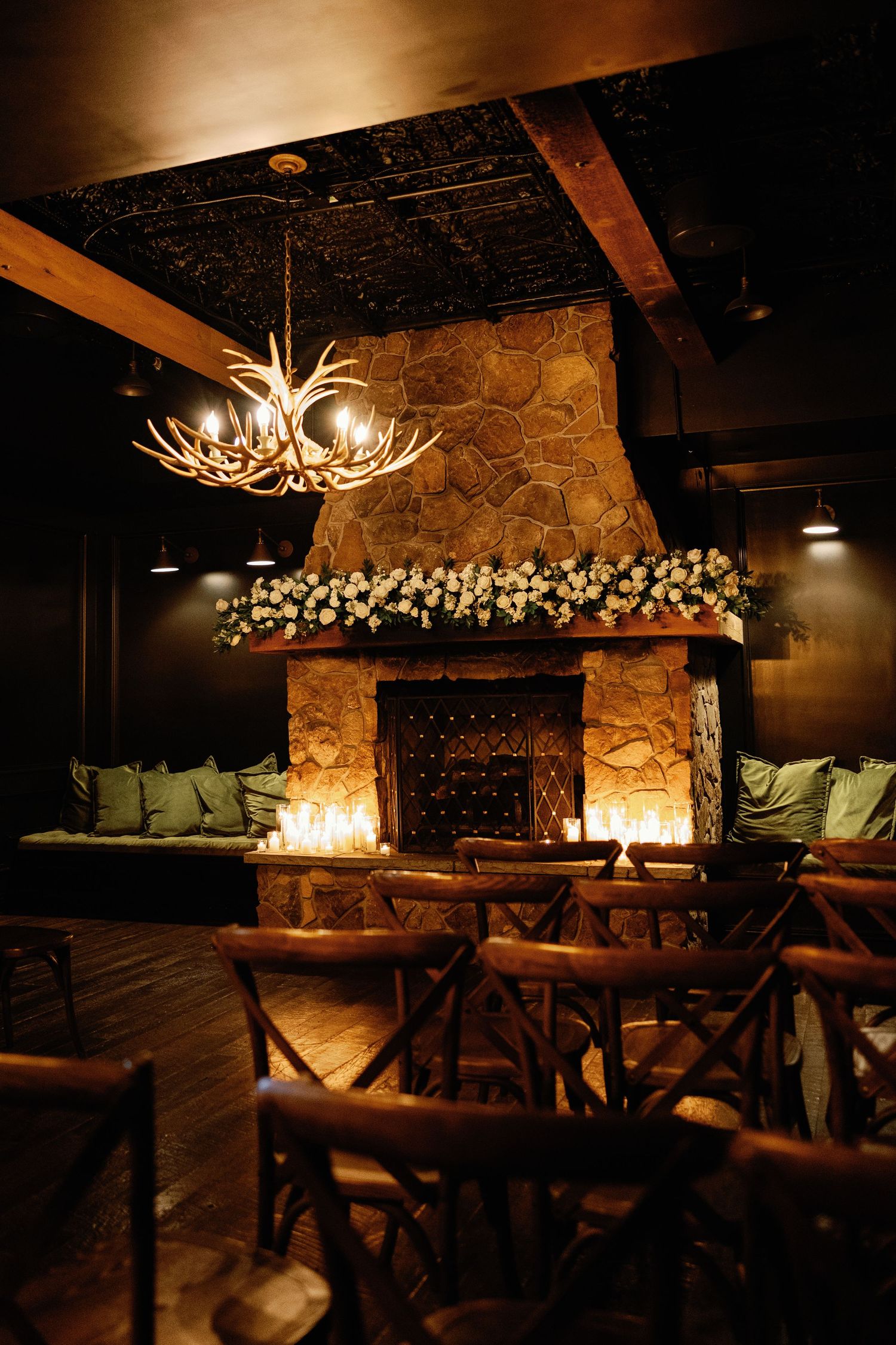Cozy wedding ceremony space with stone fireplace, white floral garland, candles, and antler chandelier.