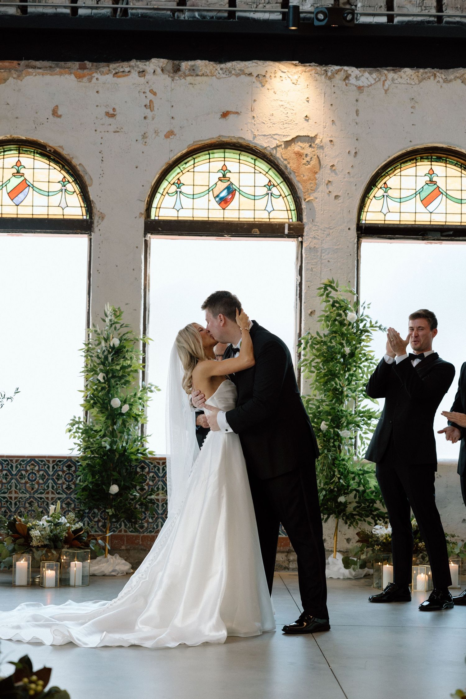 Newlyweds sharing first kiss at altar with stunning stained glass windows and greenery decor.