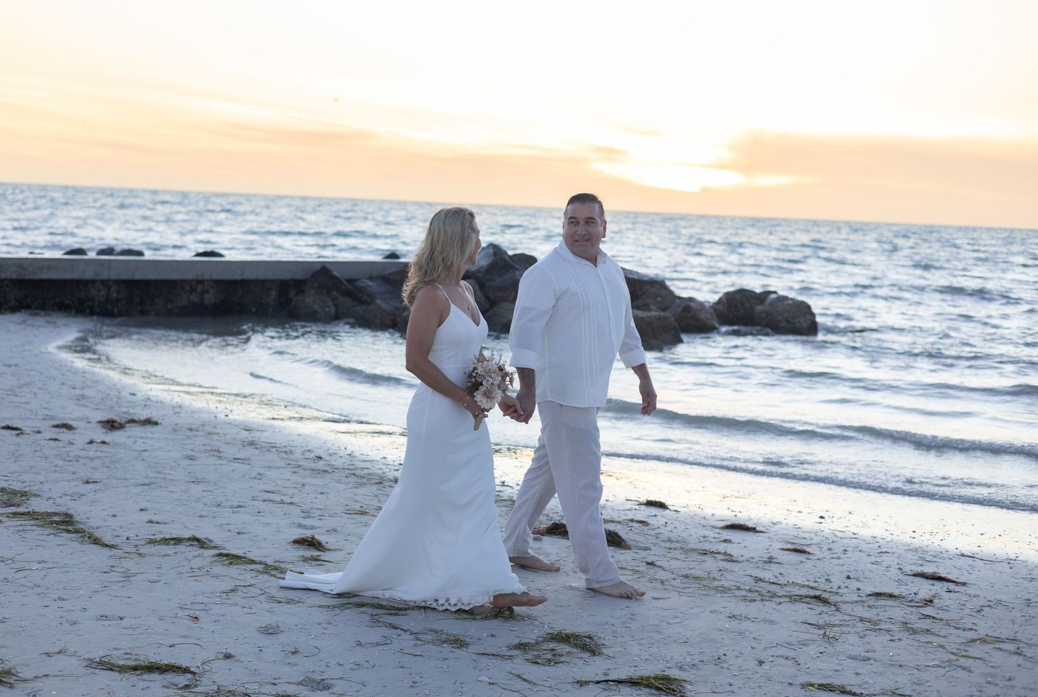 Bride and groom in white wedding attire walking hand in hand along shoreline at sunset.