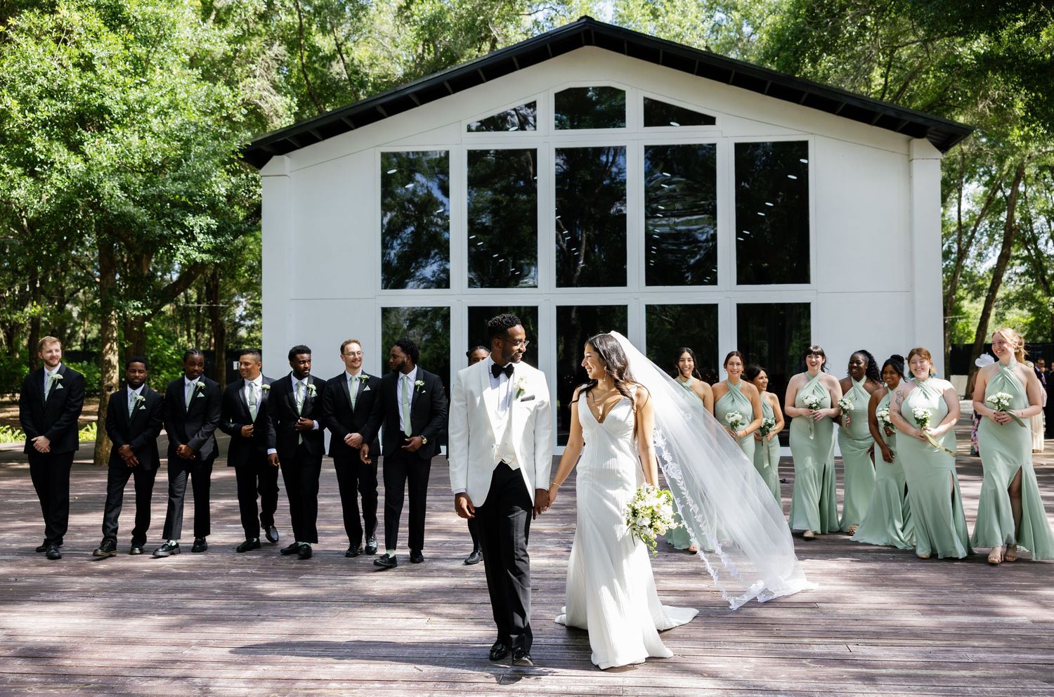 Wedding party poses outside white chapel with bridesmaids in sage green gowns and groomsmen in black suits.