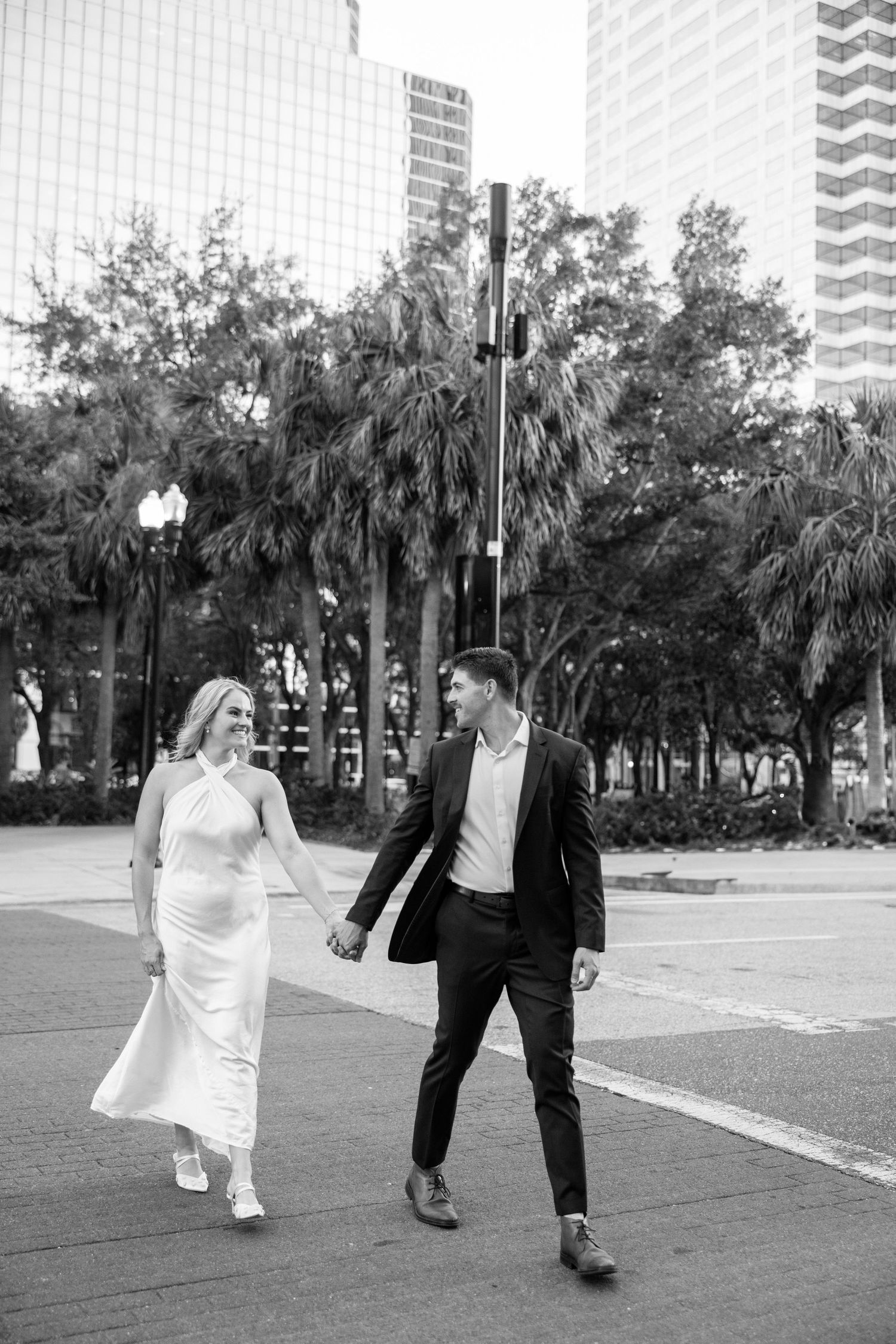 Black and white photo of engaged couple holding hands walking past palm trees in urban setting.