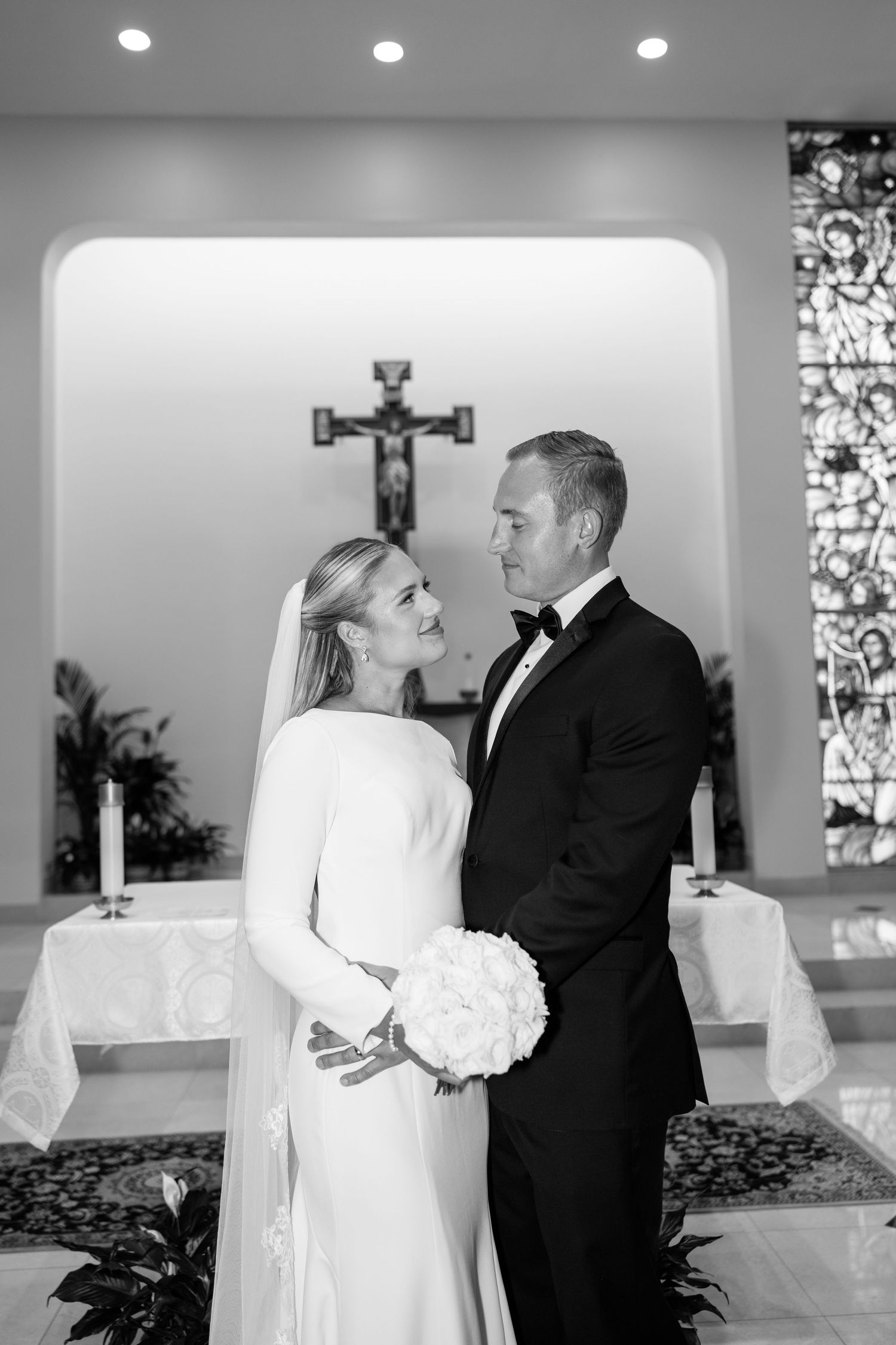 Bride and groom pose together at church altar with stained glass window in black and white wedding photo.