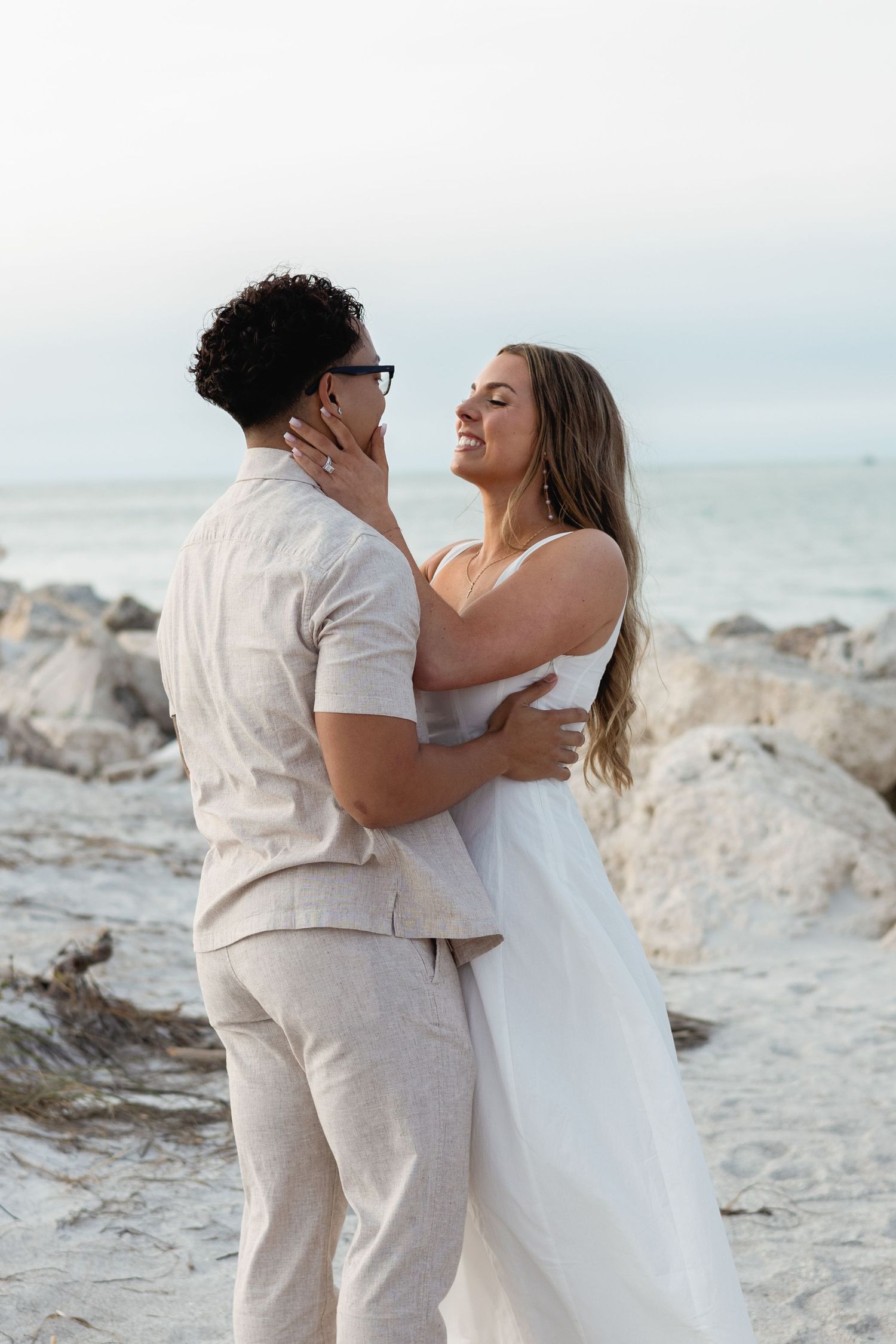 Newlyweds embrace tenderly on rocky beach with ocean in the background.