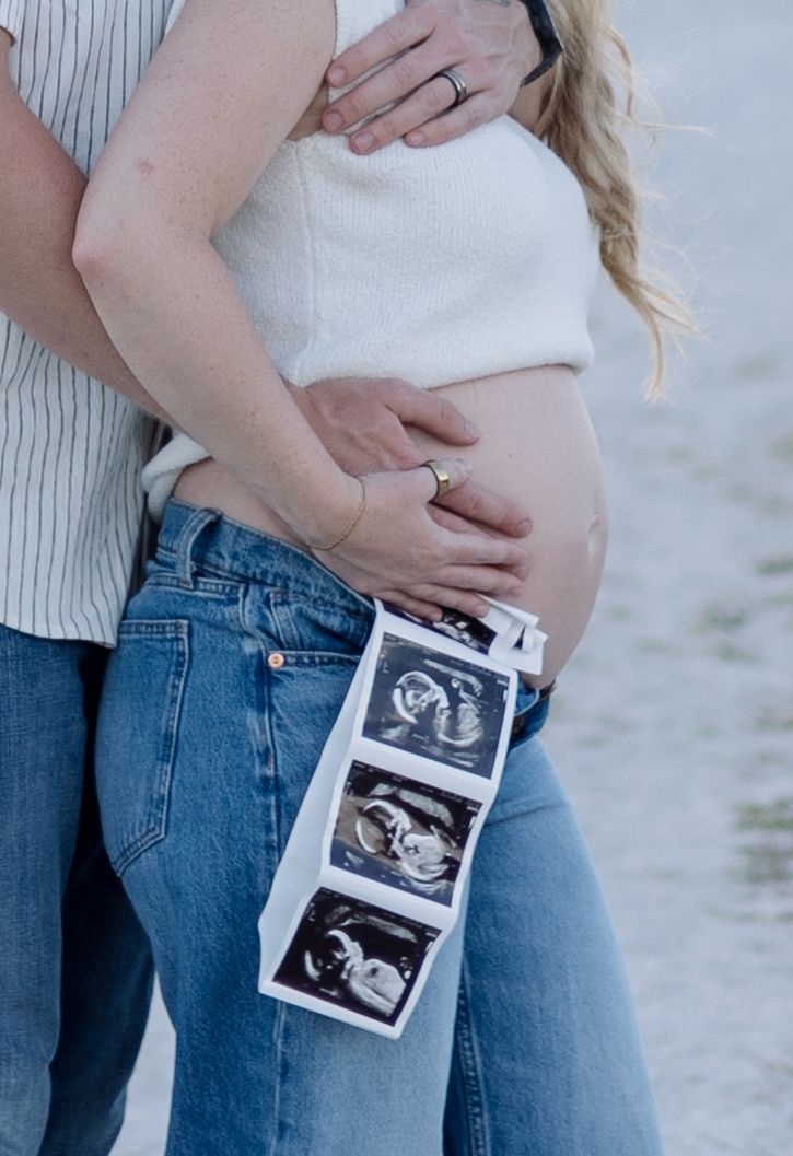 Couple holding ultrasound photos while embracing pregnant belly in casual outdoor maternity shoot.