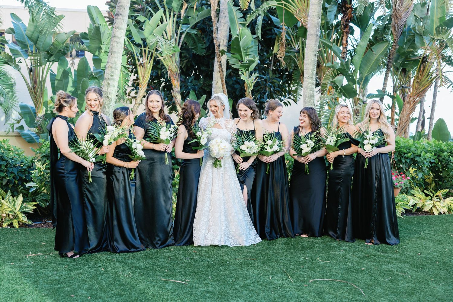 Bride in white lace gown poses with bridesmaids in black dresses holding tropical greenery bouquets outdoors among palm trees.