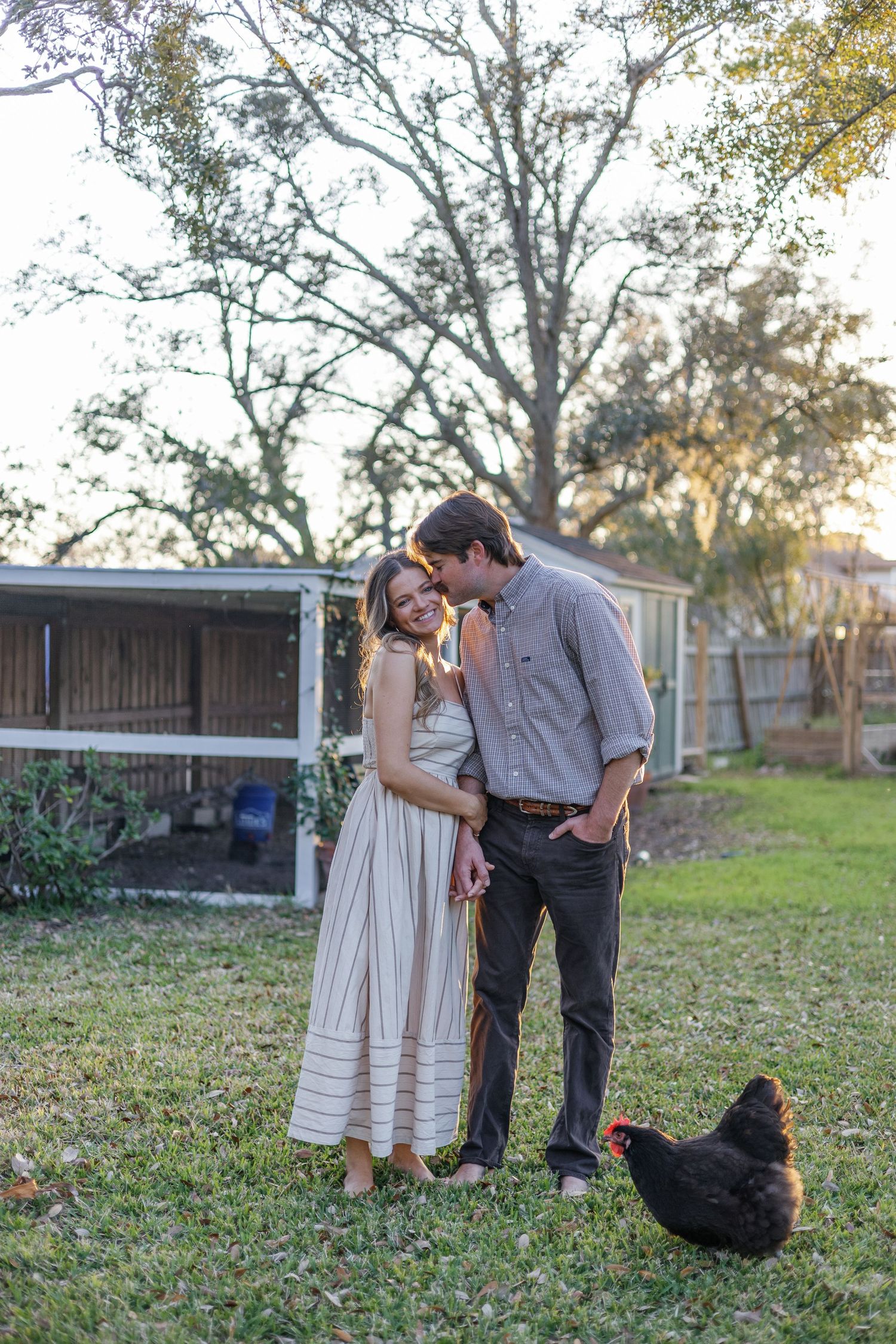 Happy couple standing in a backyard with a chicken roaming nearby at sunset.
