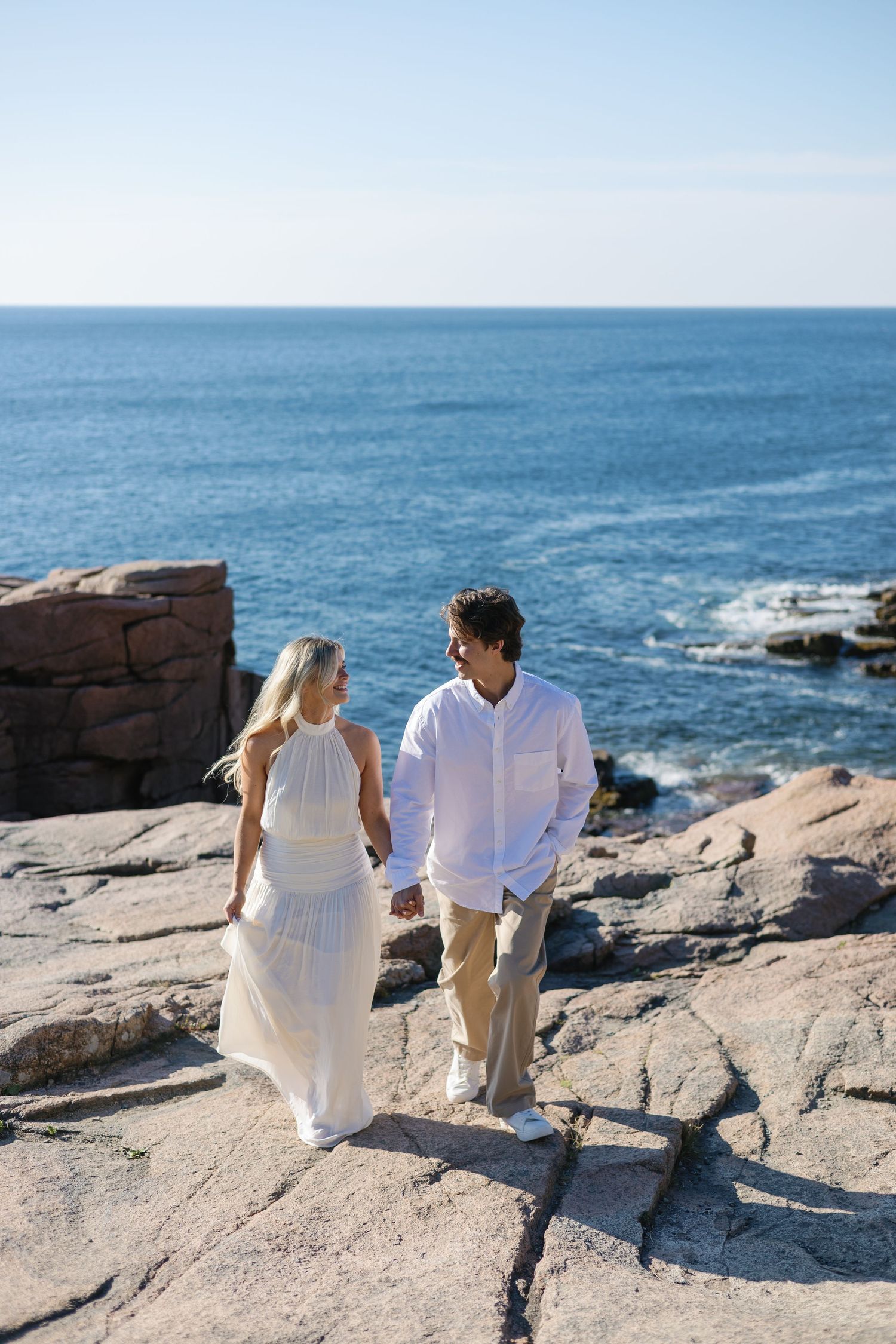 Couple holding hands while walking on rocky coastal terrain with brilliant blue ocean behind them.