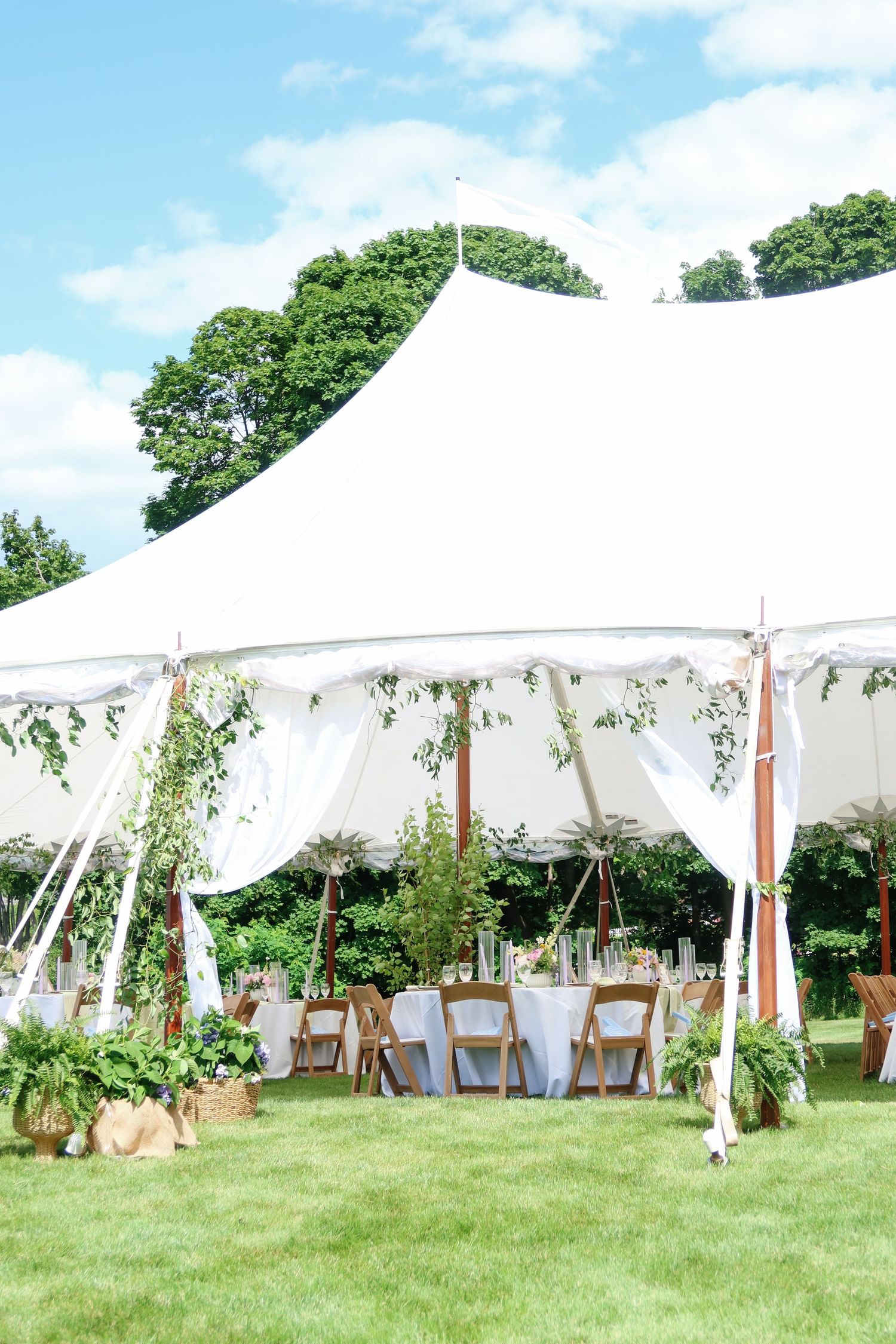 White sailcloth wedding reception tent decorated with greenery and wooden poles on sunny day with blue sky.
