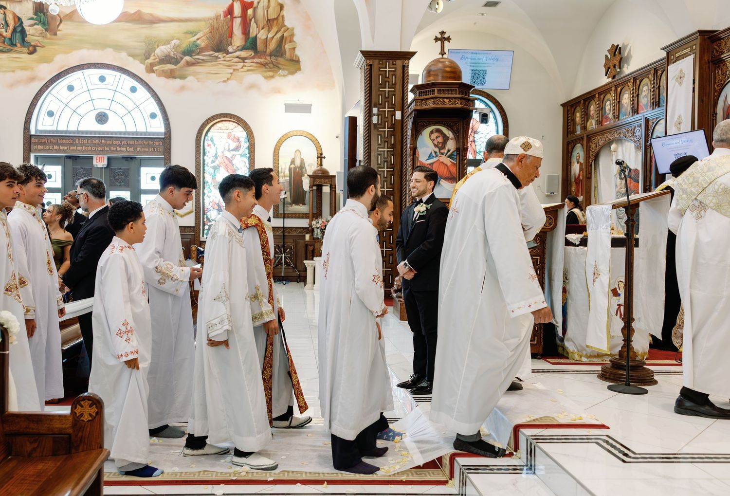 Wedding processional with altar servers in white robes walking through ornate decorated church.