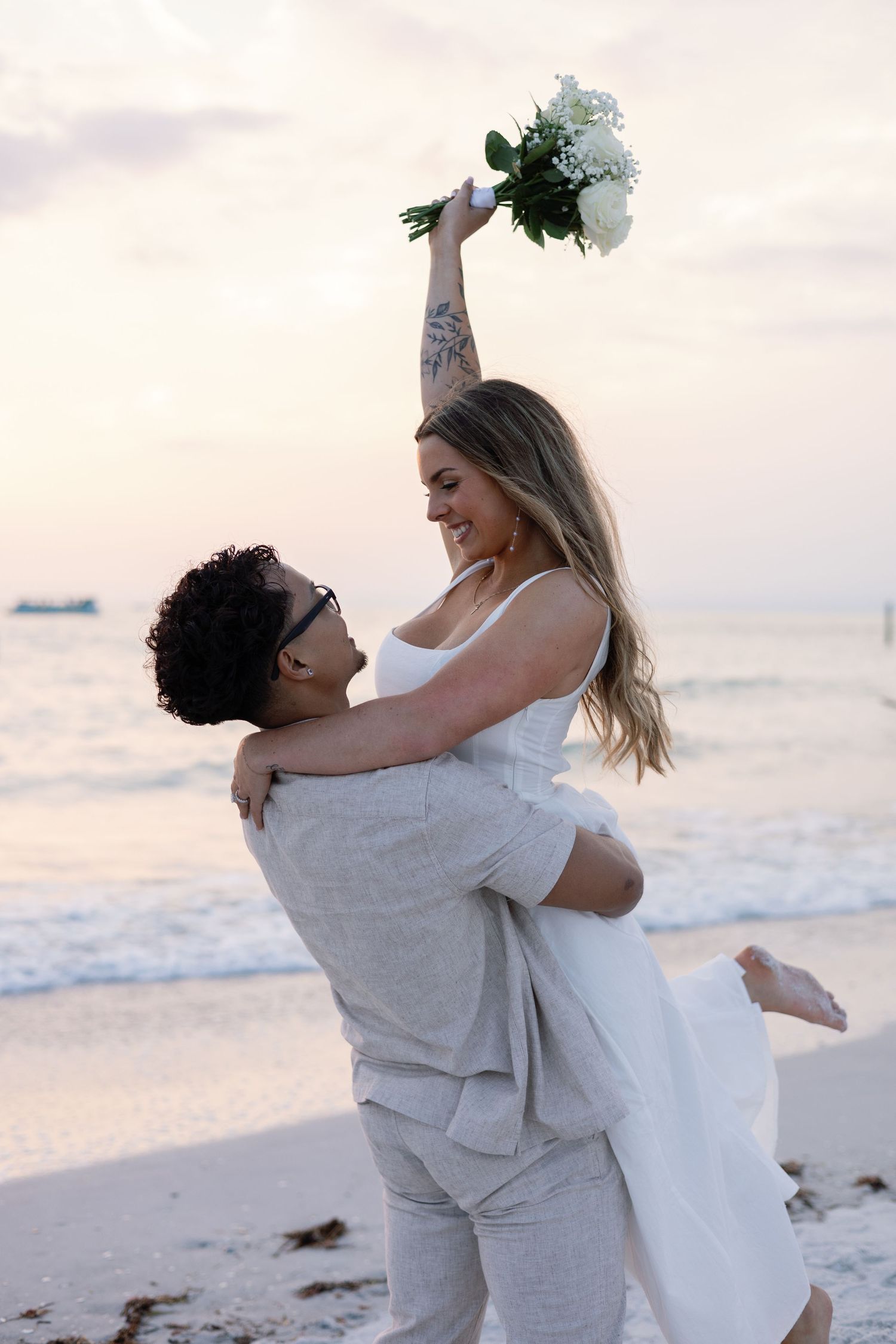 Groom lifts smiling bride holding white bouquet aloft on sandy beach at sunset.