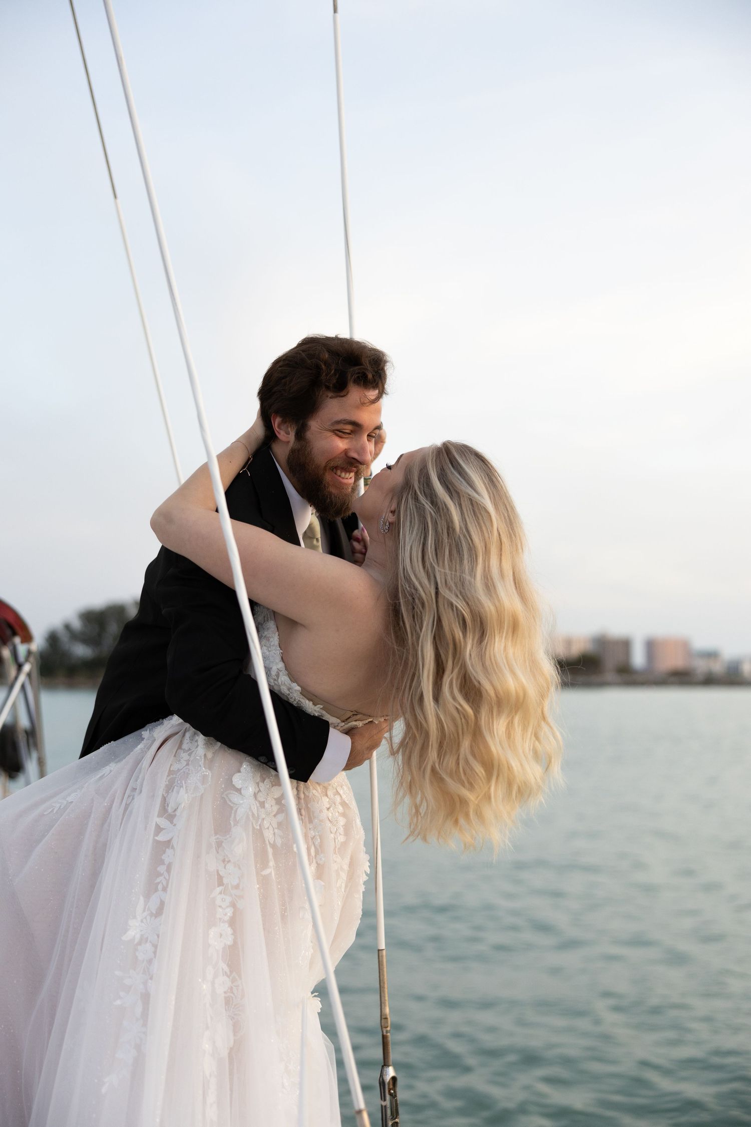 Bride and groom share romantic moment embracing on sailboat with water and city skyline behind them.