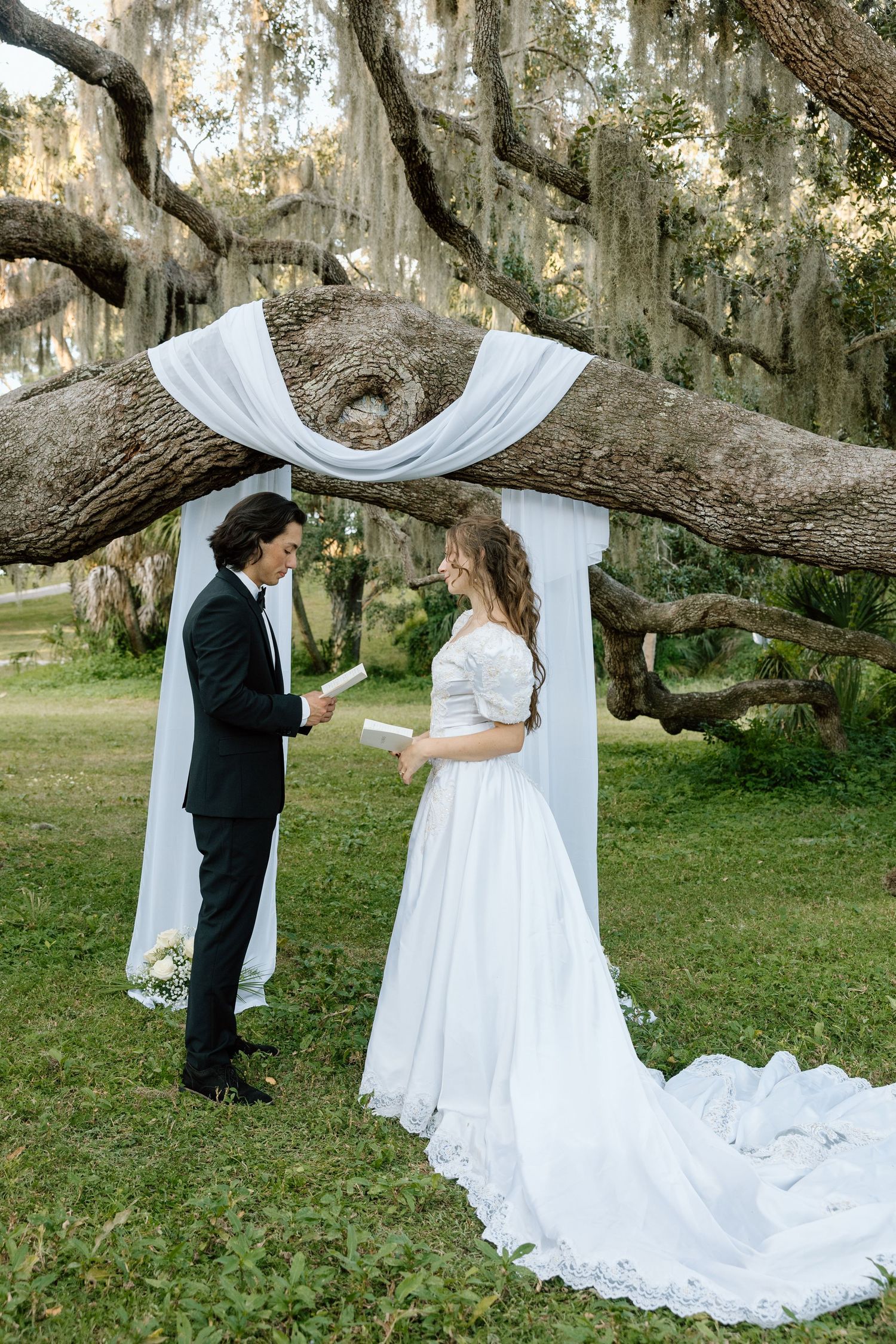 Groom reads vows to his bride beneath a white draped arch framed by a massive moss-covered oak tree in a lush green setting.