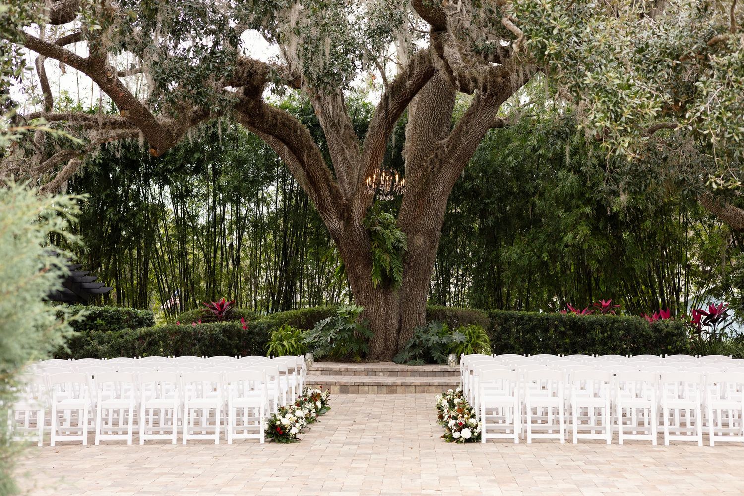 Outdoor wedding ceremony setup with white chairs facing a grand oak tree altar in a garden.