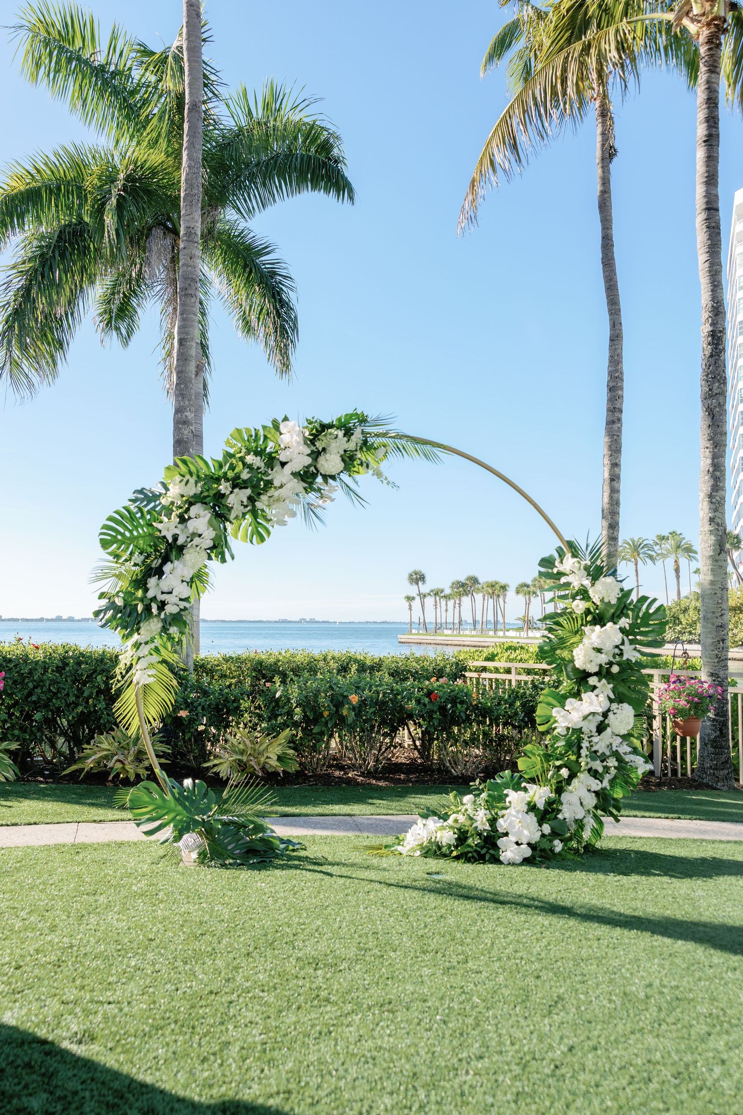 Circular wedding arch adorned with white flowers and tropical greenery on a lush lawn with palm trees and ocean views.