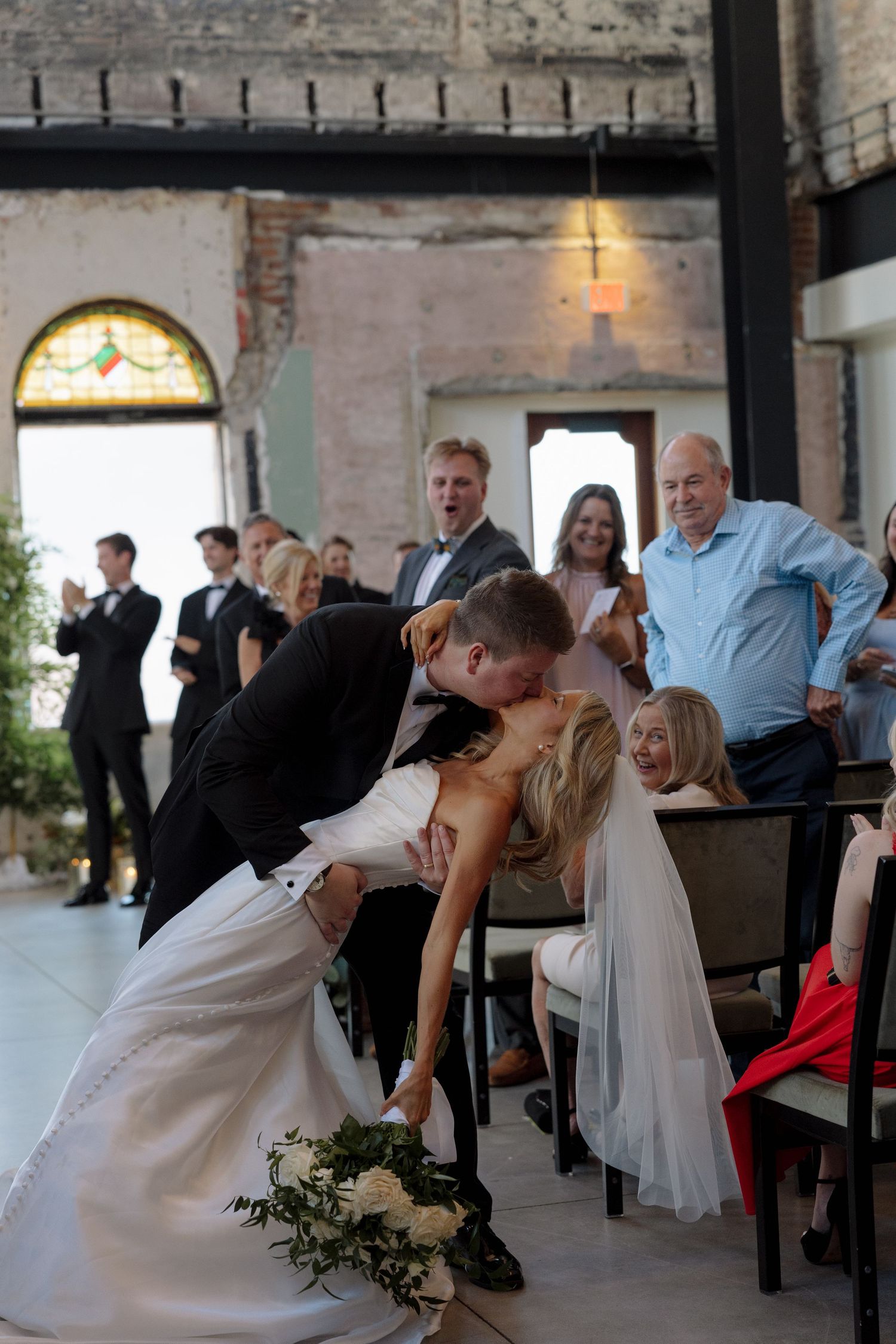 Bride and groom share a romantic dip kiss at wedding ceremony while guests cheer in rustic brick venue.