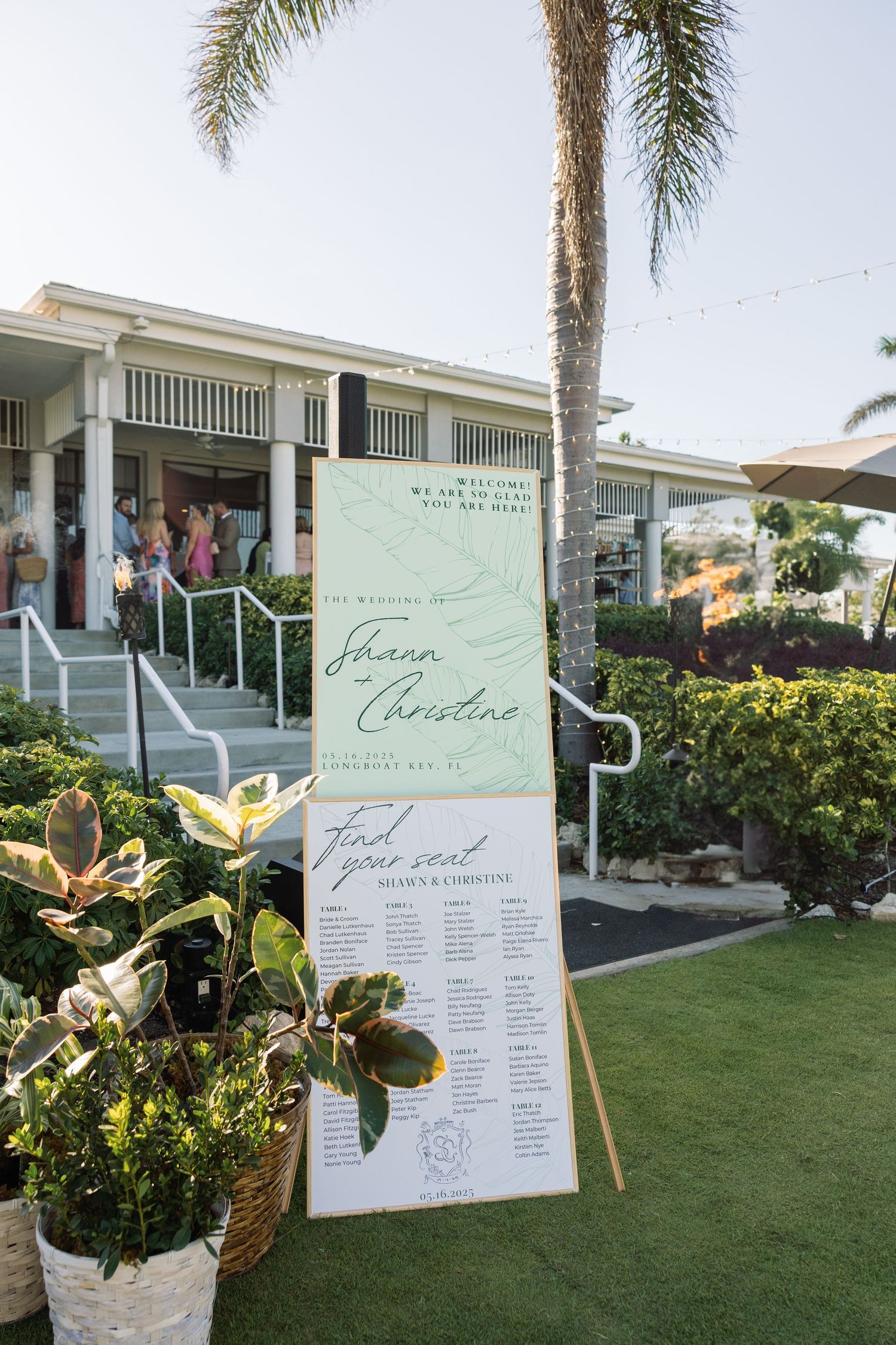 Elegant green and white wedding seating chart sign displayed outdoors at tropical venue with palm trees and string lights.
