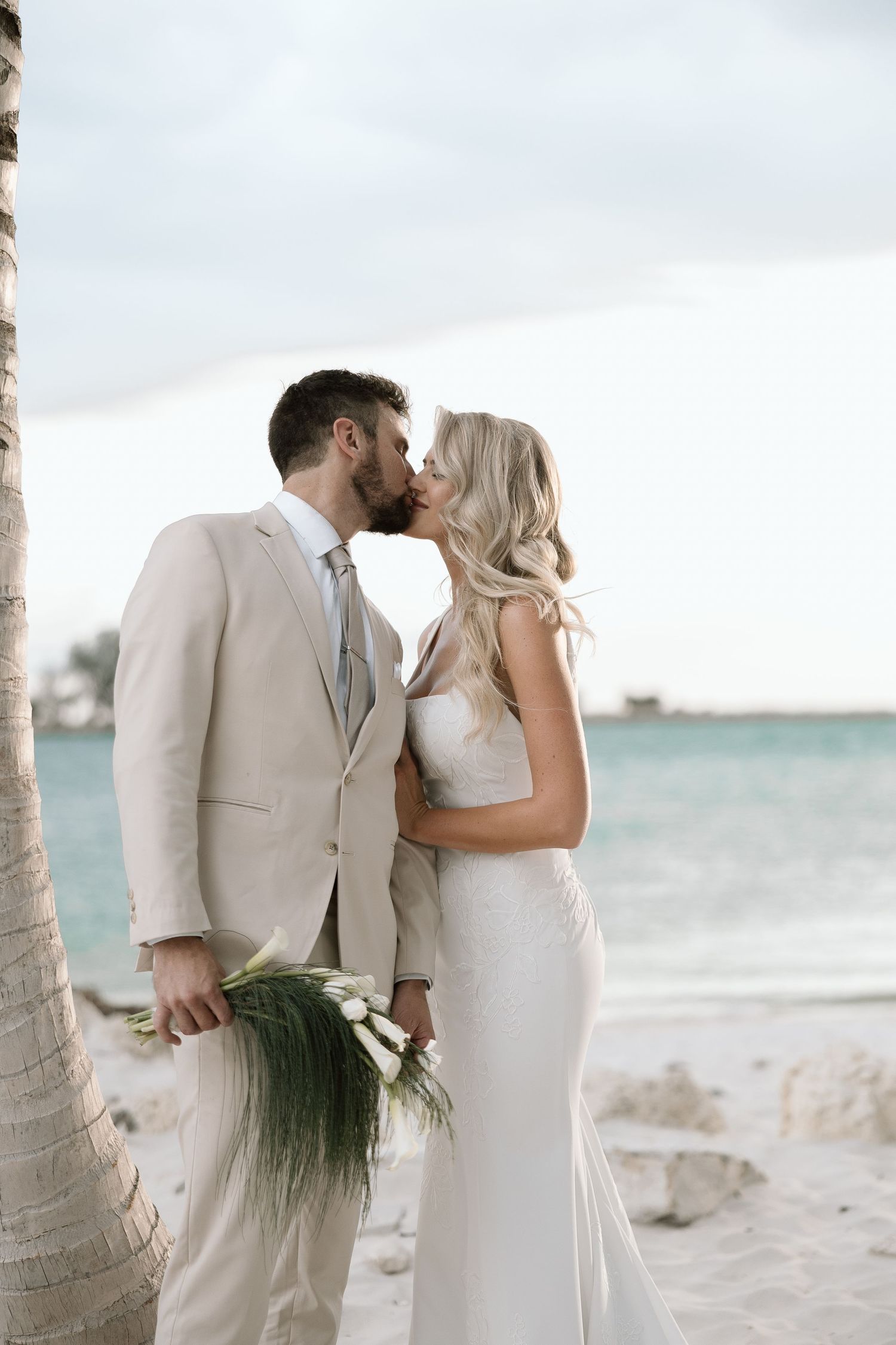 Bride and groom sharing tender kiss by palm tree on sandy beach, groom holding tropical greenery bridal bouquet.