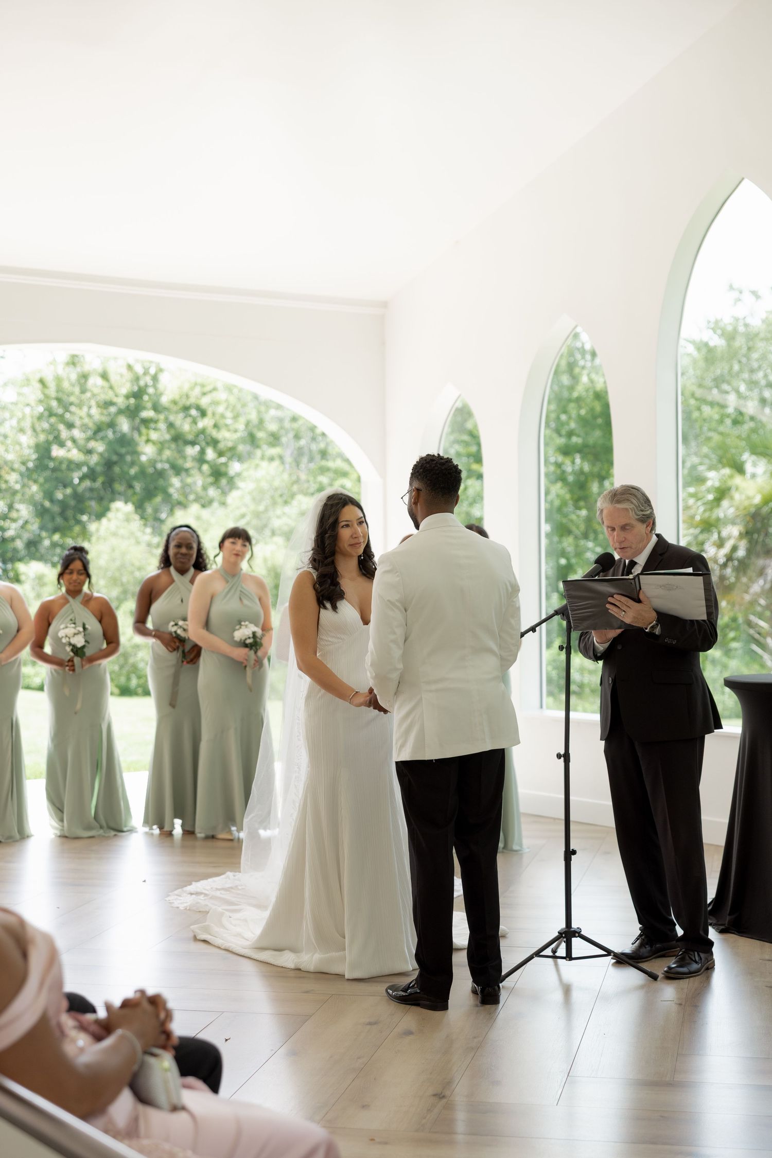 Bride and groom hold hands as officiant reads vows with bridesmaids in sage green dresses.