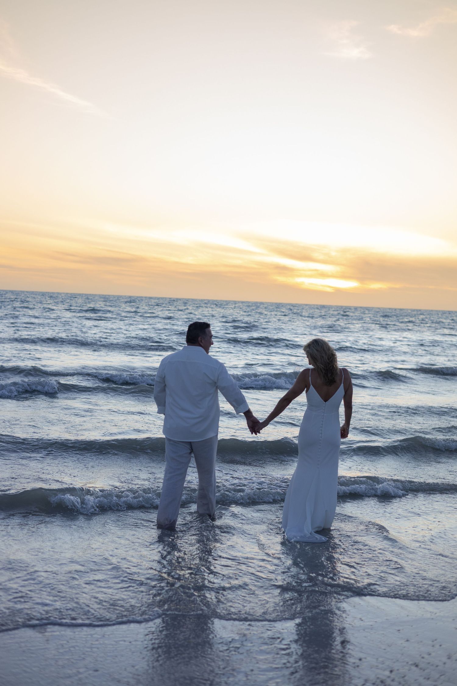 Newlywed couple holding hands standing in ocean waves watching breathtaking golden beach sunset.