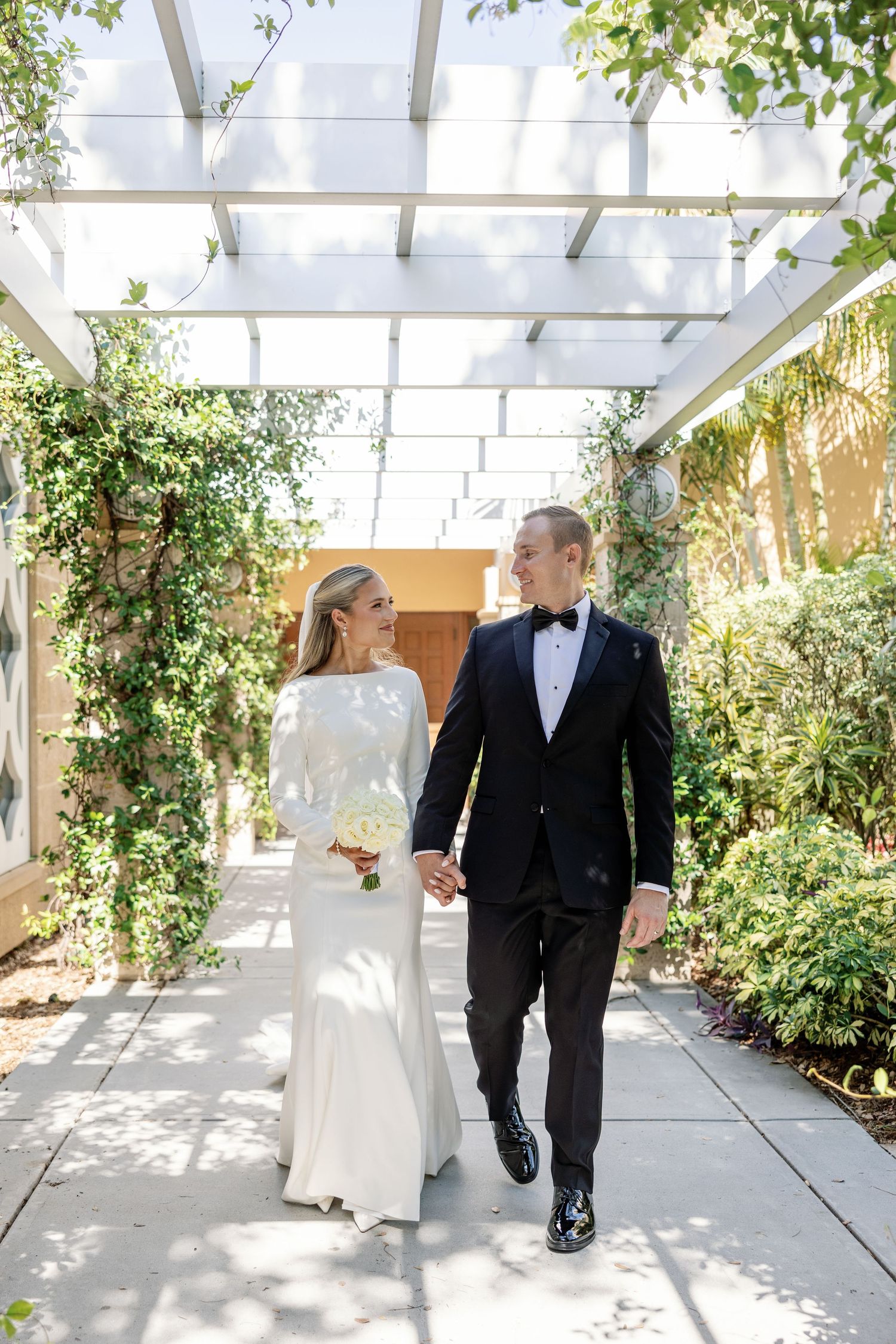 Bride and groom walk hand in hand under ivy-covered pergola in bright outdoor wedding portrait.