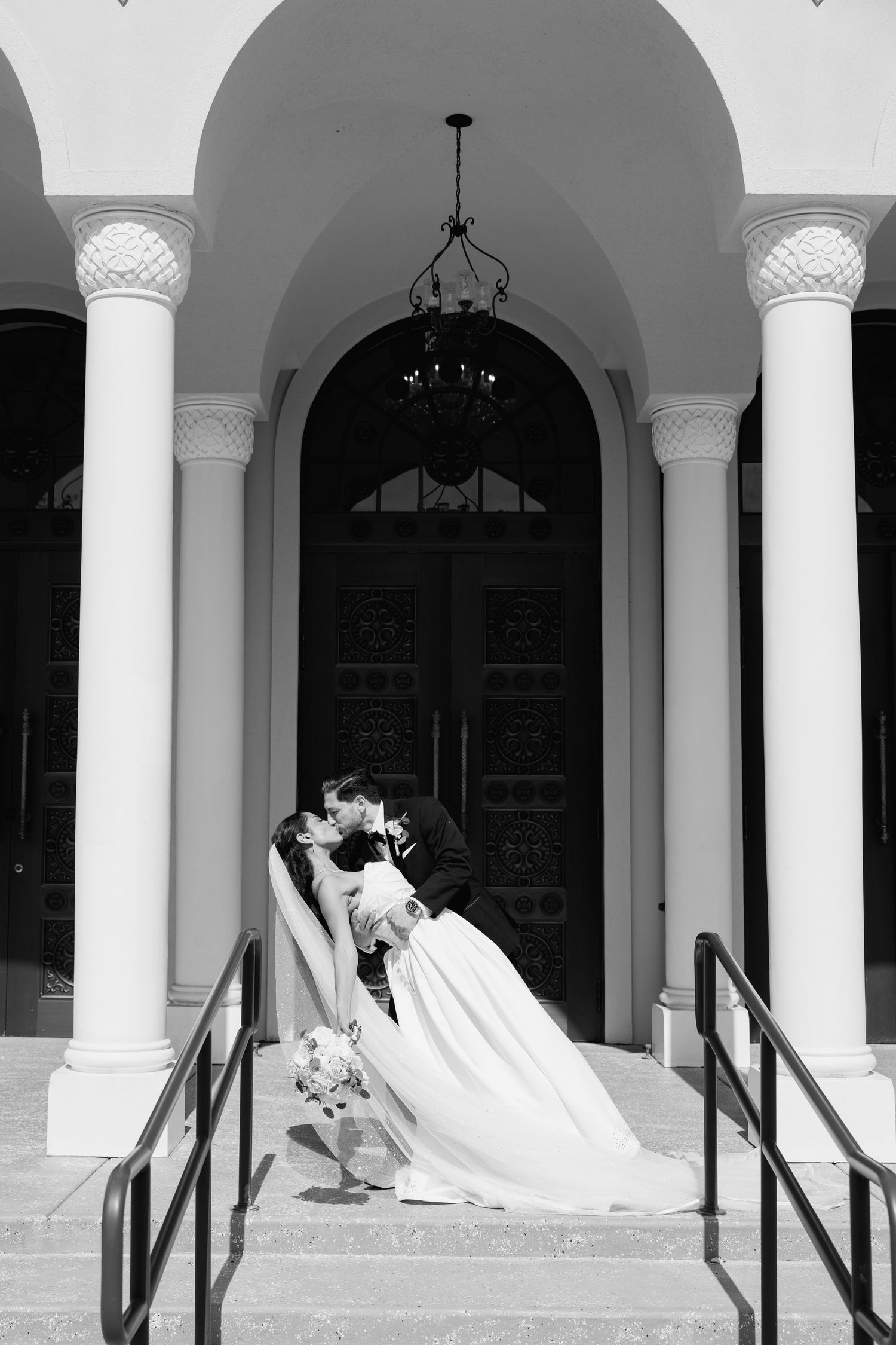 Bride and groom share romantic dip kiss on church steps in elegant black and white wedding portrait.