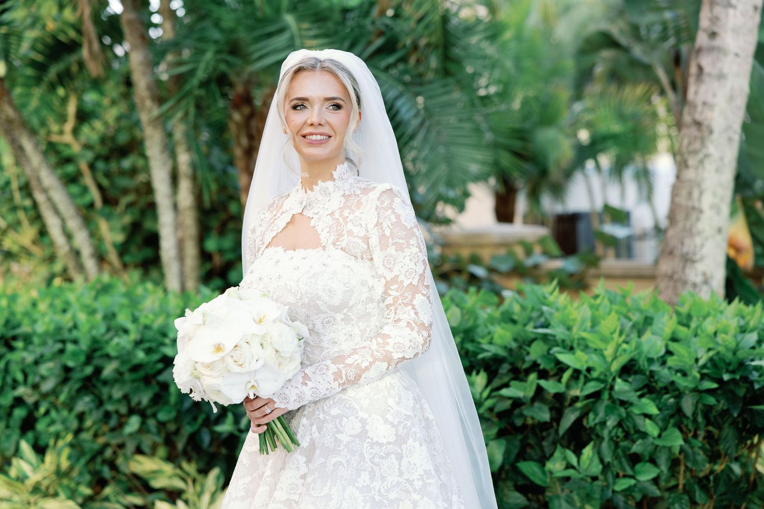 Bride in lace long-sleeve wedding gown and veil holding white bouquet, standing outdoors among tropical greenery.