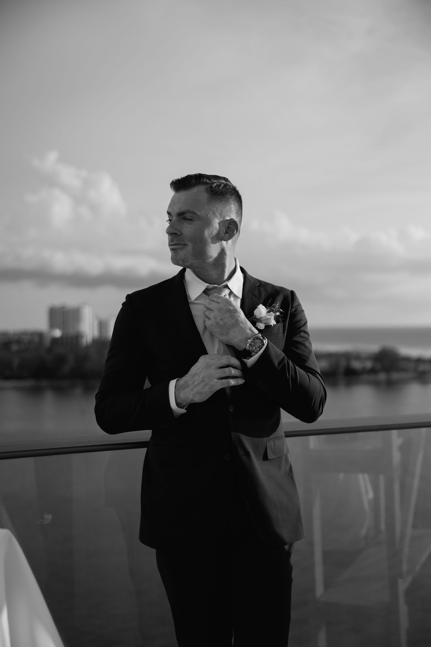 Black and white portrait of smiling groom in black suit adjusting jacket on waterfront balcony.