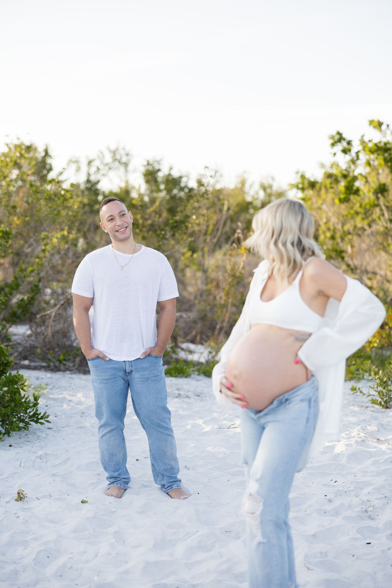 Smiling man in white tee watches pregnant partner display her bare baby bump on sandy beach.