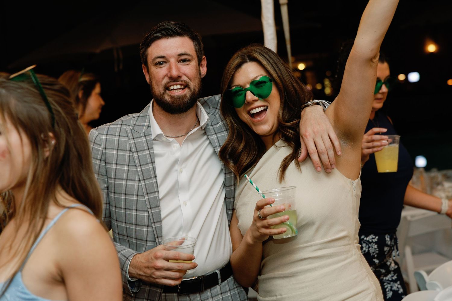 Couple celebrating at wedding reception, woman wearing green heart sunglasses raising arm joyfully while holding drinks under tent.