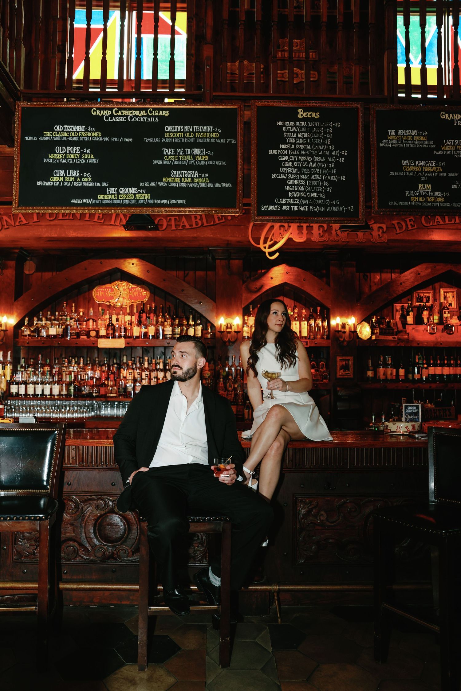 Elegant couple sits apart at a gothic-style bar with stained glass windows and extensive liquor shelves.