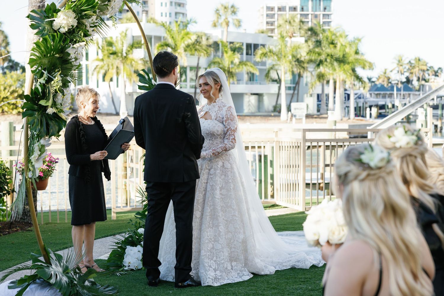 Outdoor waterfront wedding ceremony with bride in lace gown, groom in black suit, and officiant under floral arch.