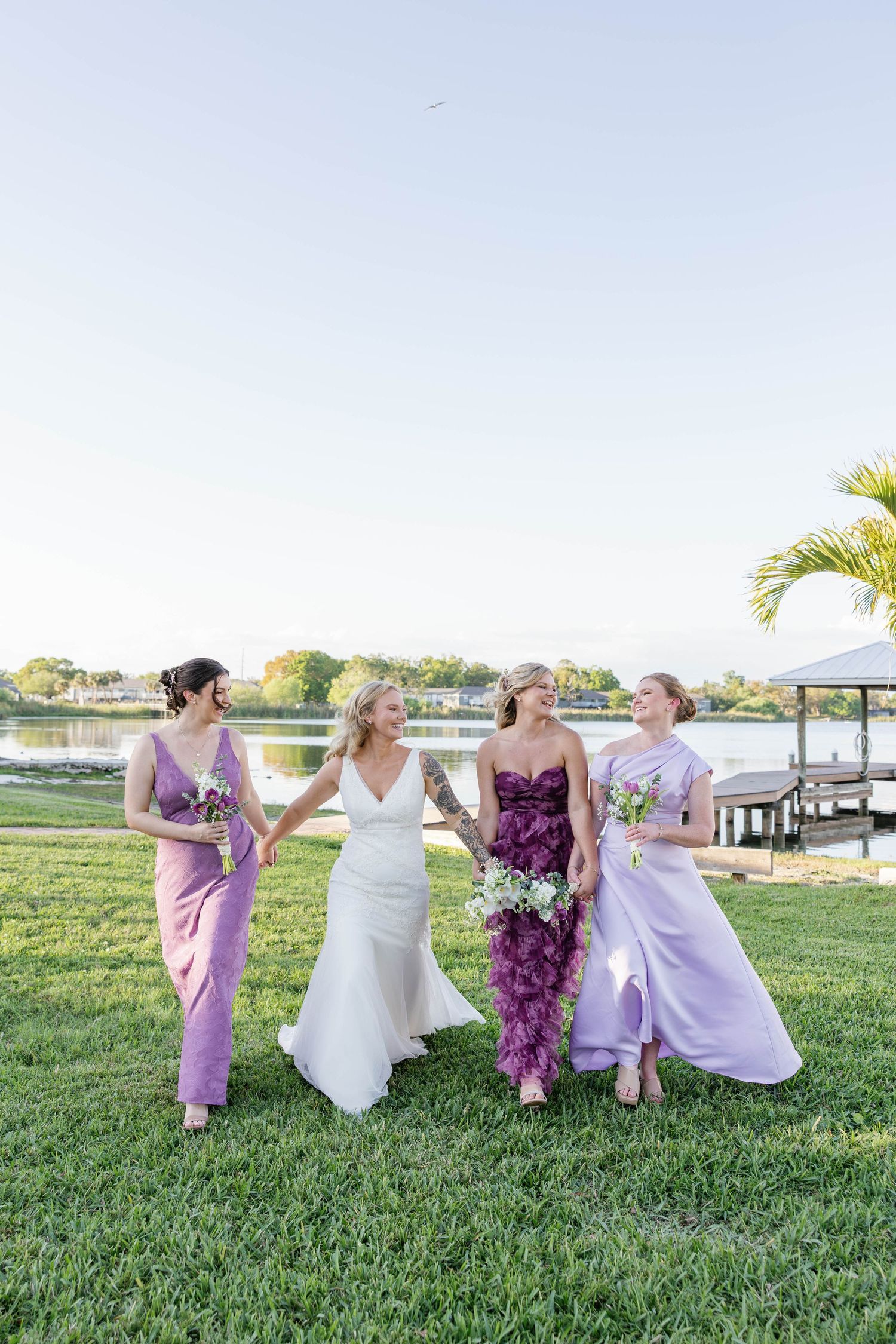 Bride in white gown walks with three bridesmaids in purple and lavender dresses along a waterfront lawn.