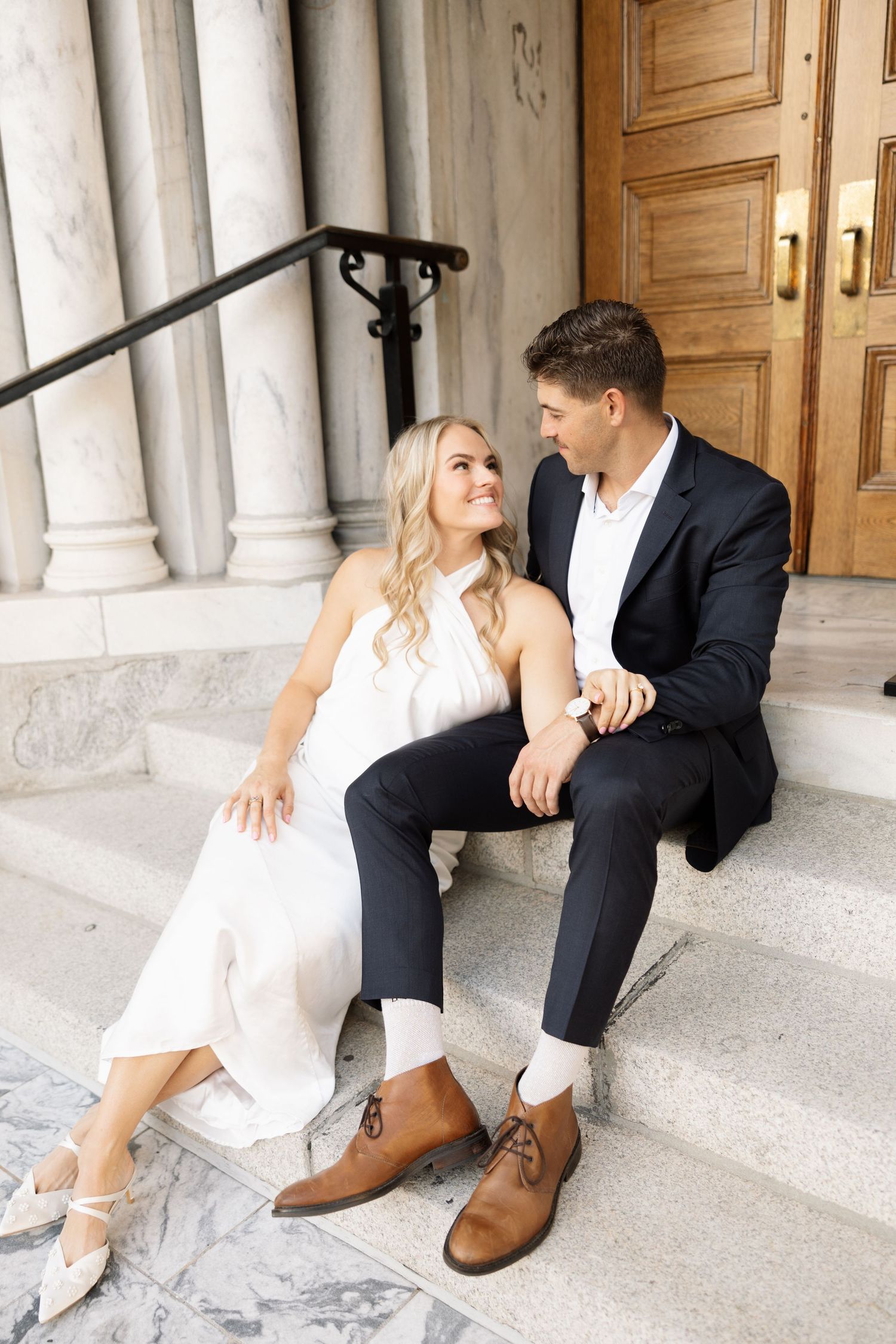 Couple in wedding attire sit on marble steps of grand building smiling at each other.